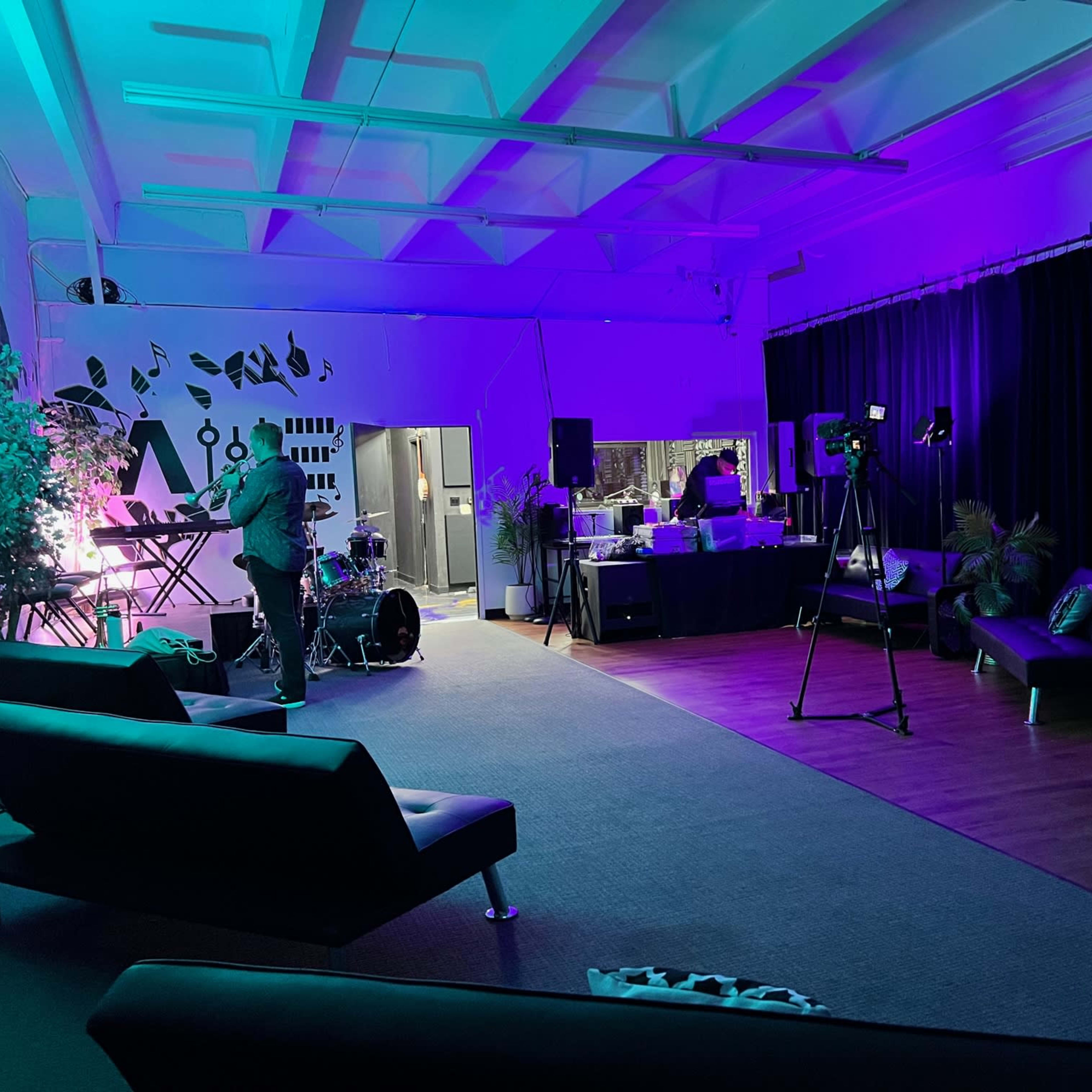 A lively interior space set up for a music event, featuring a stage with instruments, lighting, and seating areas.