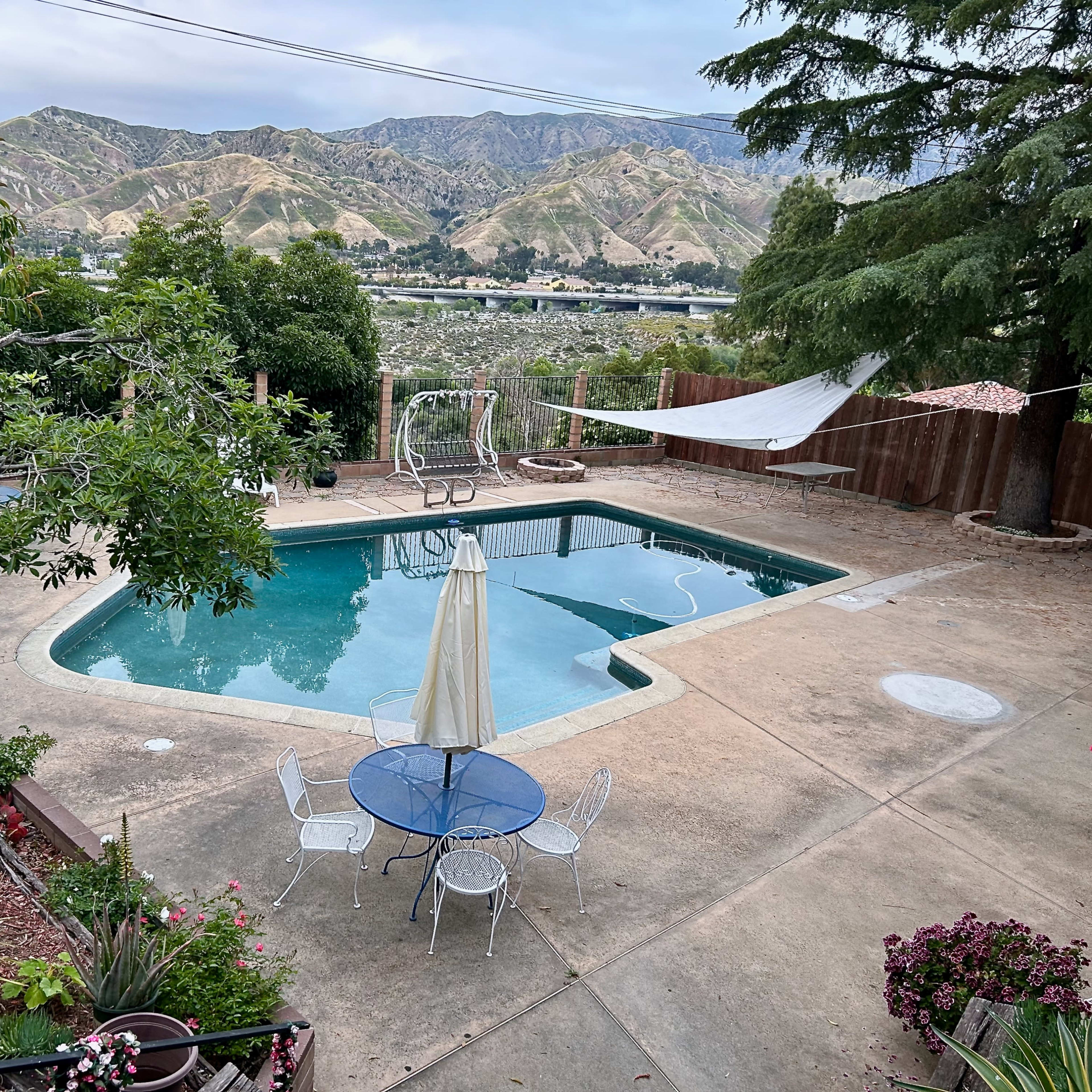 A backyard with a swimming pool, a dining table with chairs, and mountainous scenery in the background.