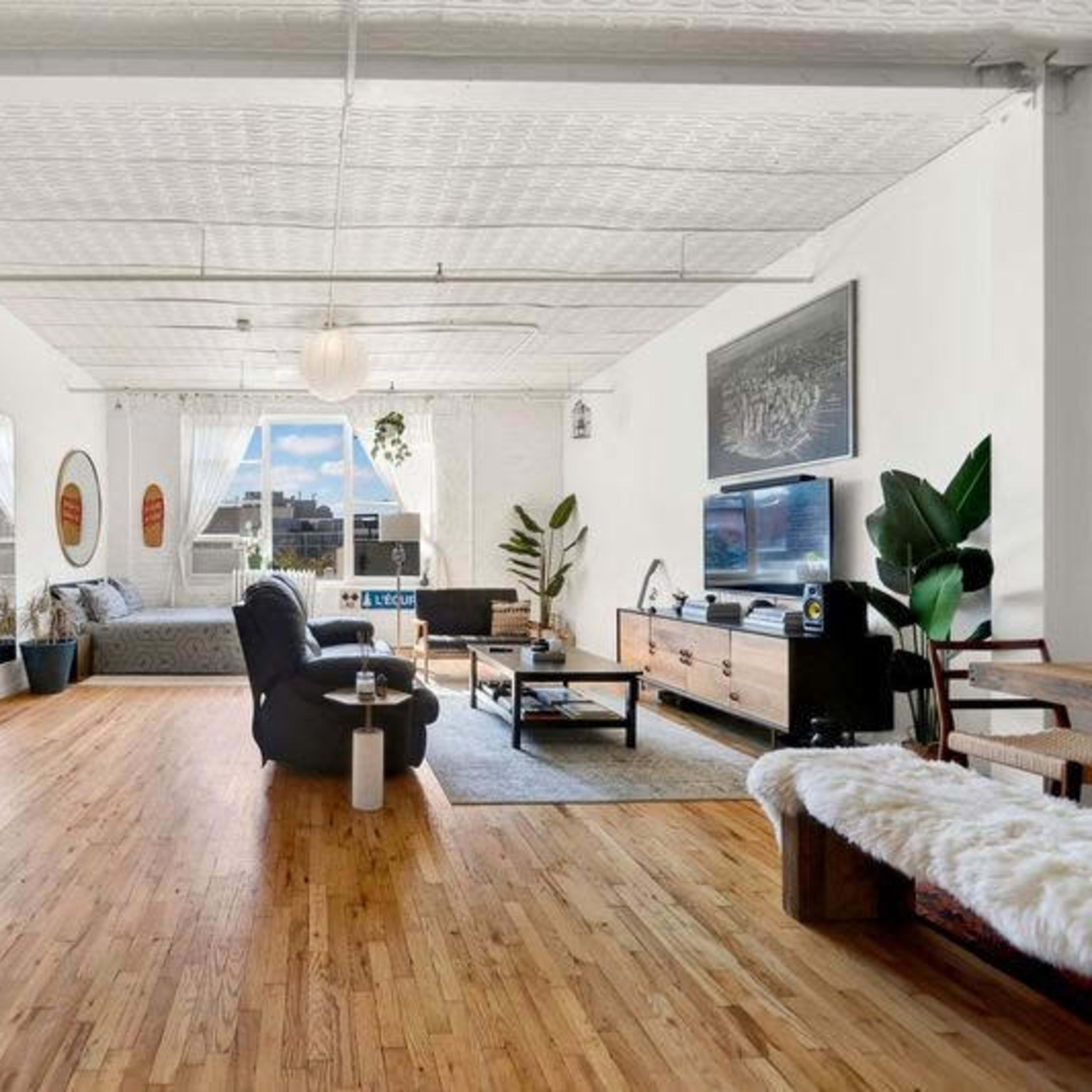 A bright, spacious living area with wooden flooring, large windows, a modern sofa, and a wooden media console against the wall.