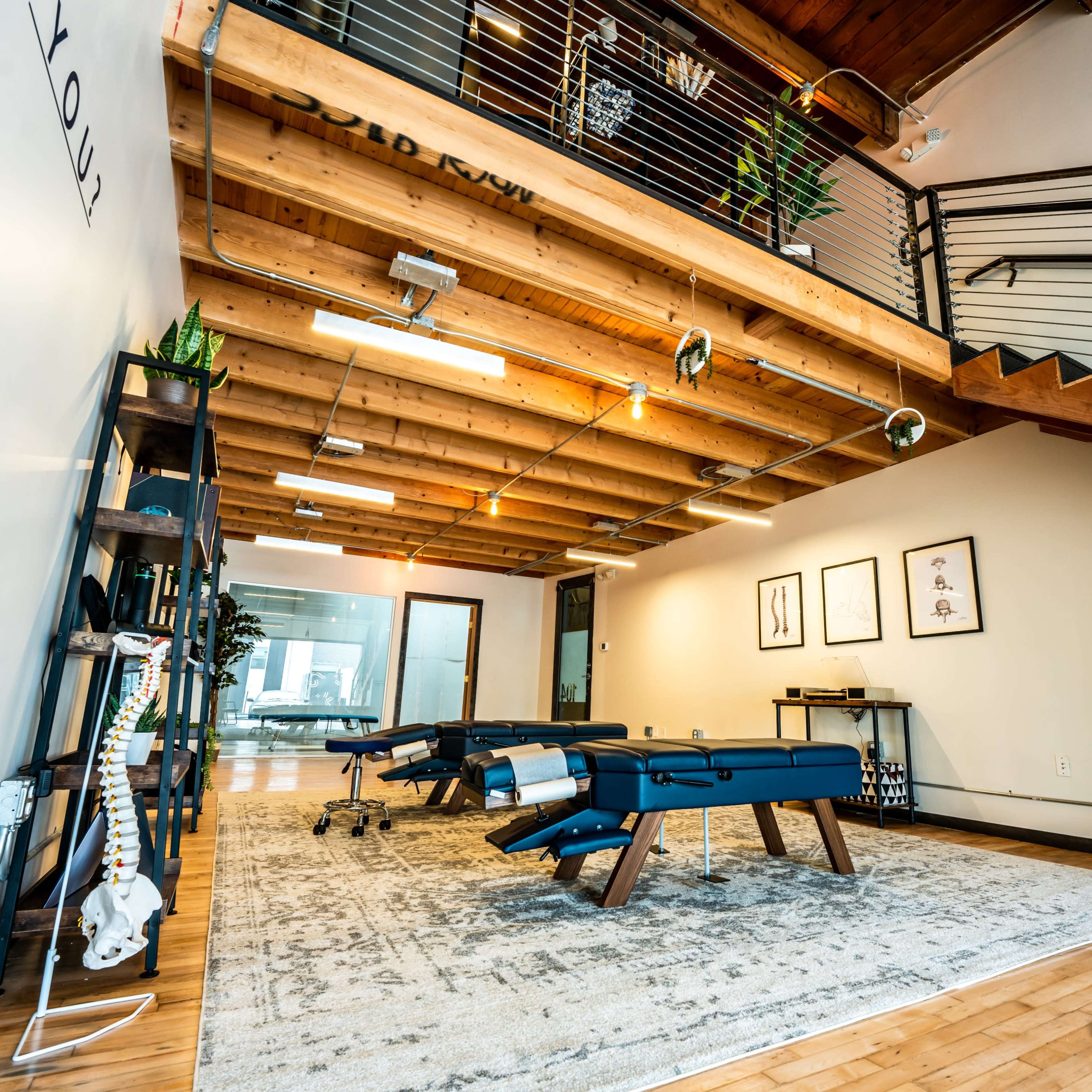 A spacious interior with wooden beams and a carpeted area containing treatment tables and decorative plants, leading to a staircase.
