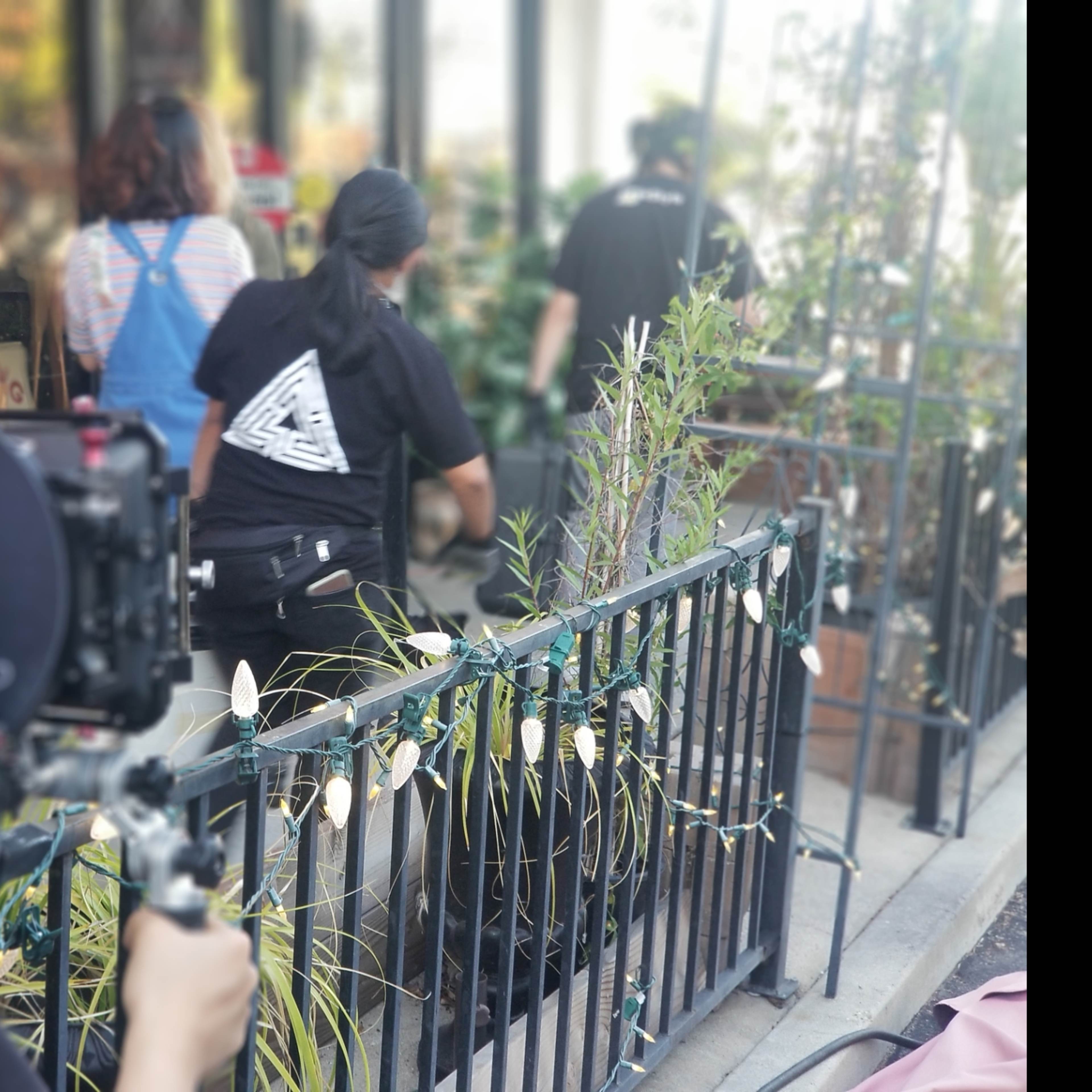 A film crew sets up equipment while two individuals walk past a railing adorned with plants at a filming location.