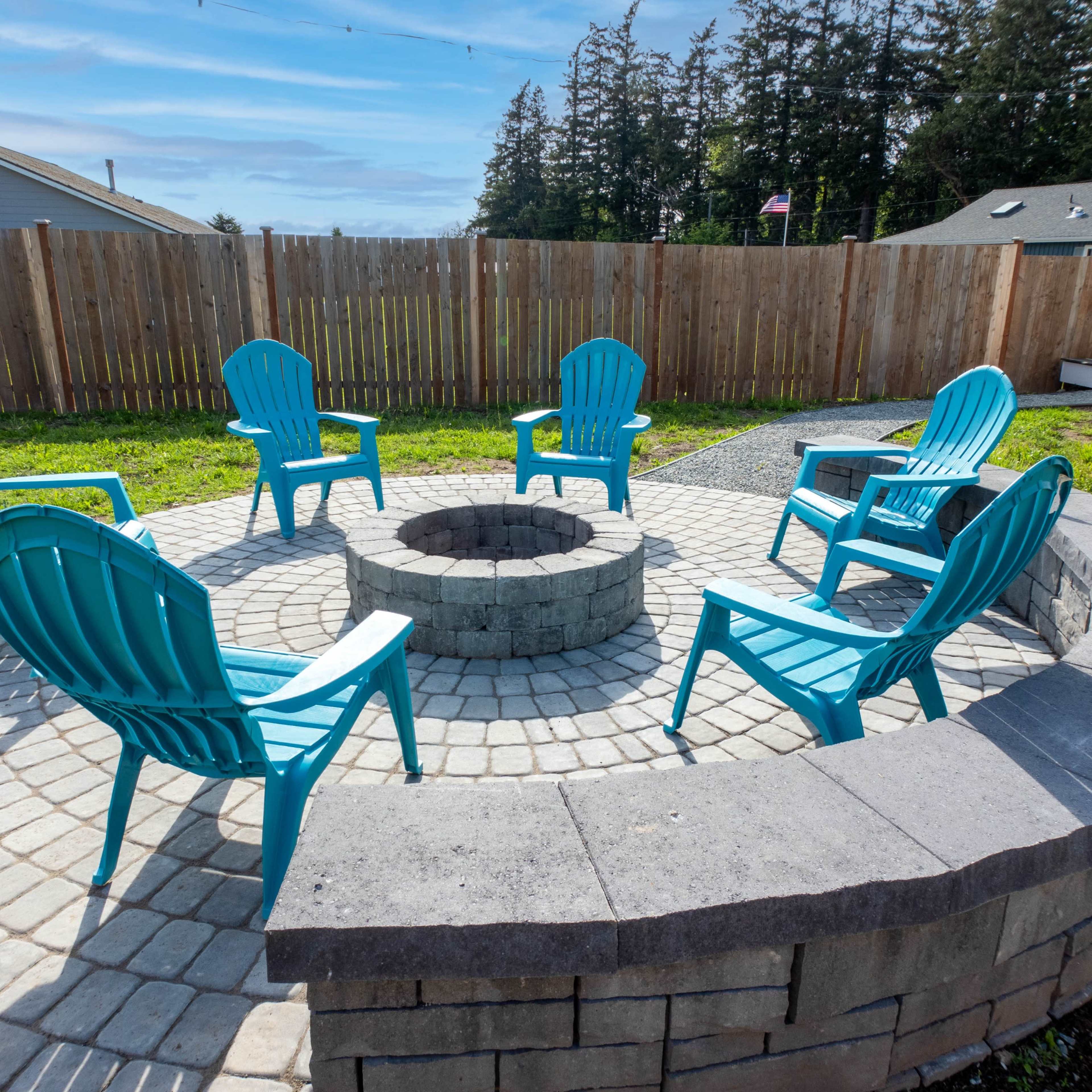 A circular stone fire pit surrounded by six blue plastic chairs in an outdoor setting.