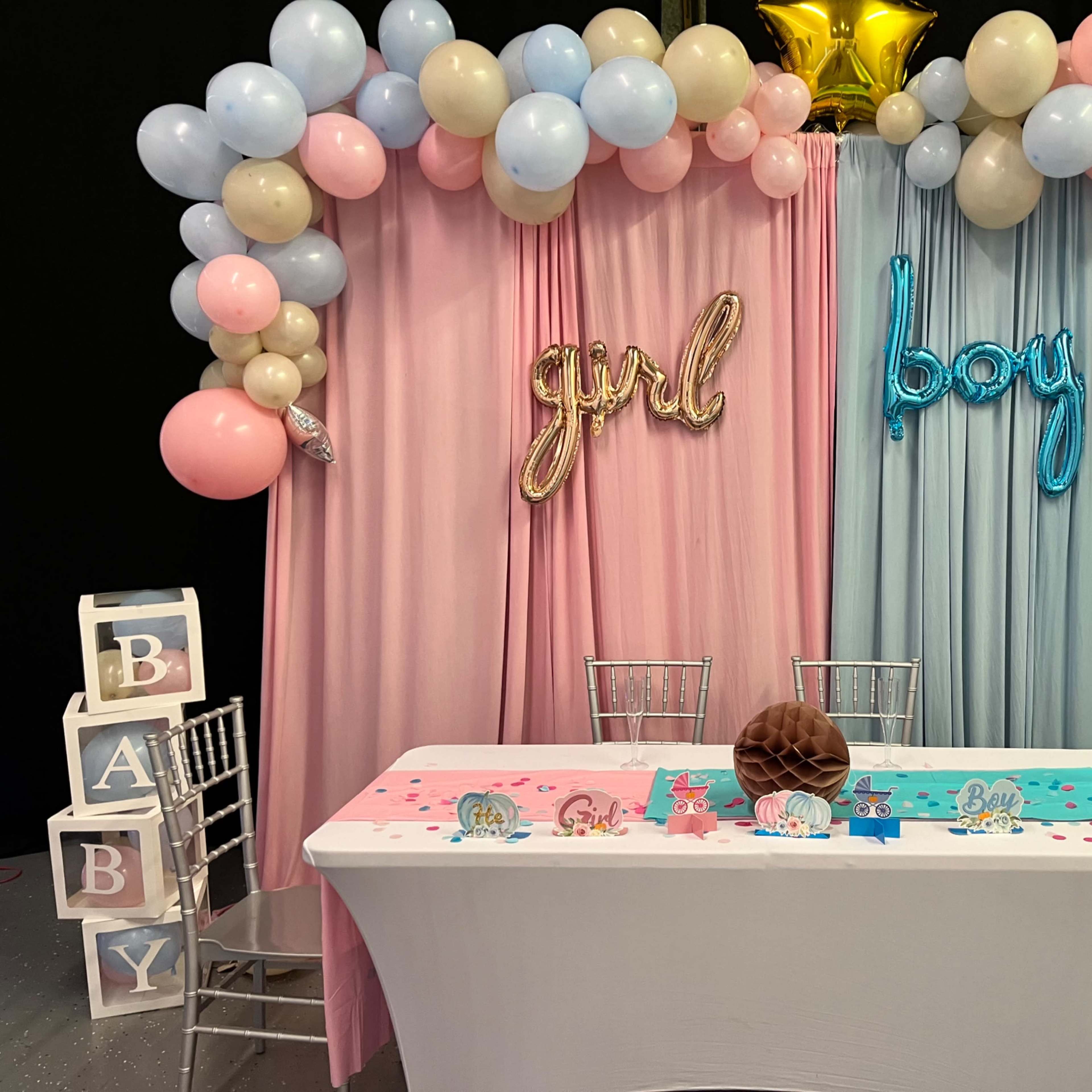 A decorated table for a baby shower, featuring pink and blue balloons, a "girl" and "boy" sign, and a table draped with pastel colors.