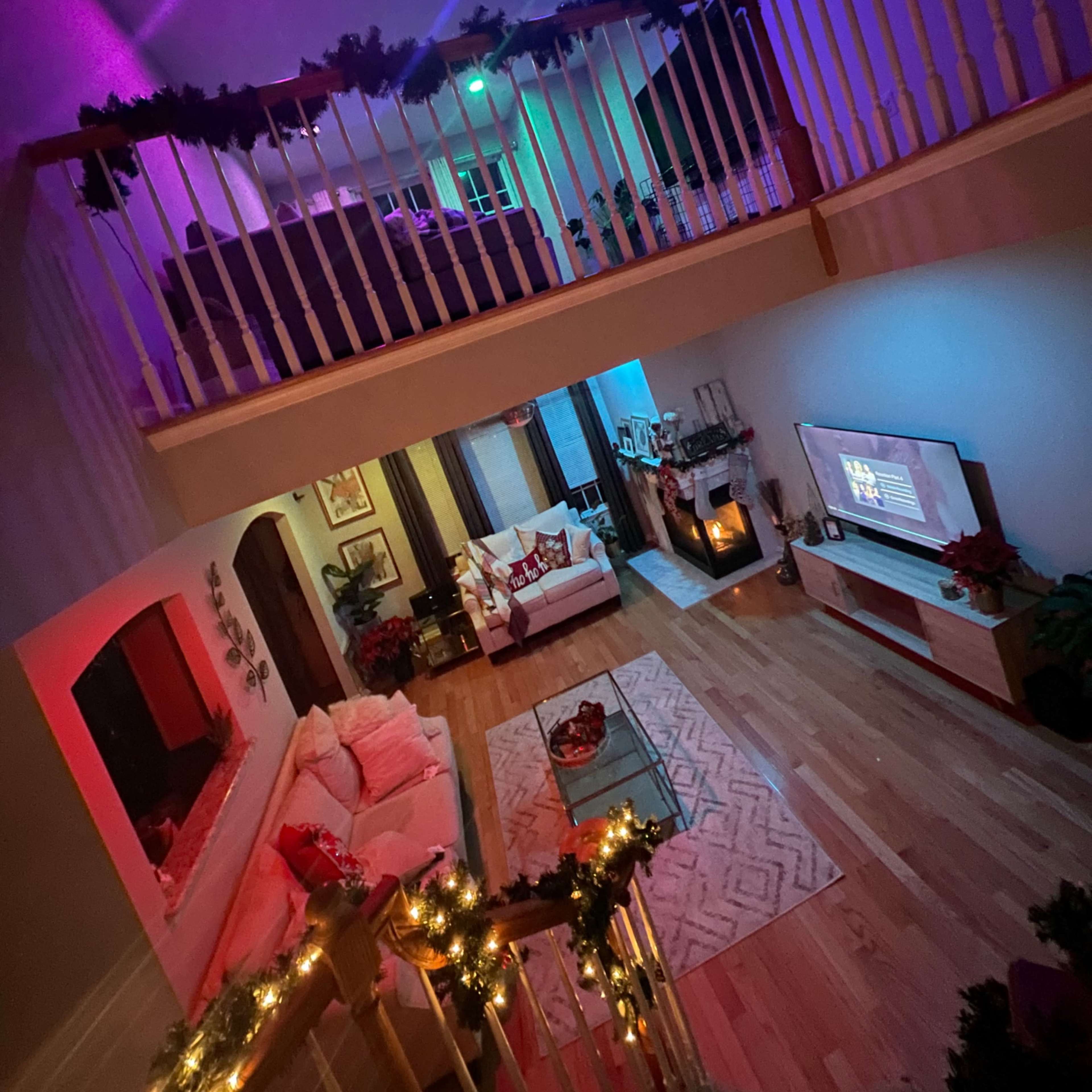 A two-story living room decorated with colorful lights and garlands, featuring a modern sofa, a television, and a fireplace.