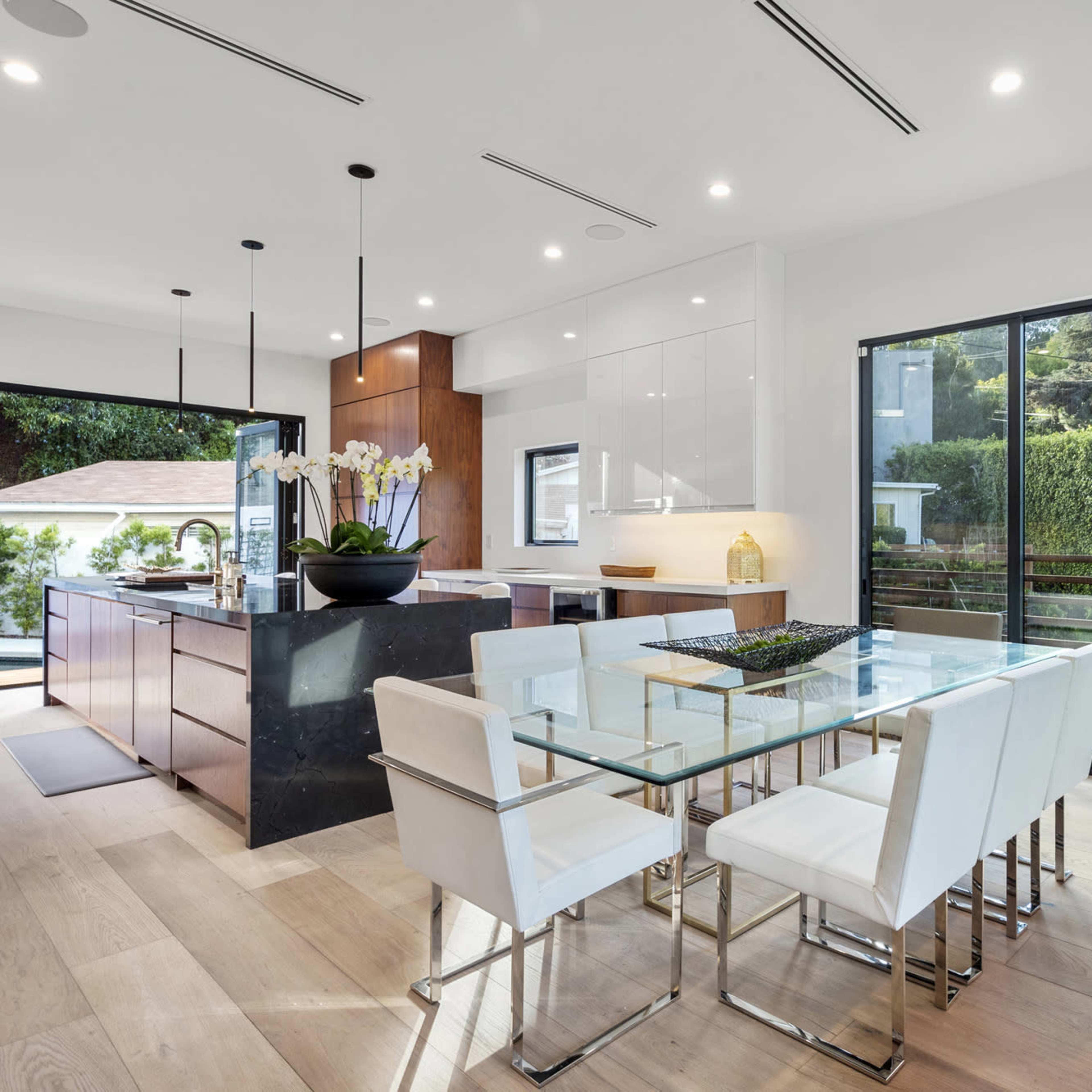 A modern kitchen and dining area features a glass dining table surrounded by white chairs, with an island and sleek cabinetry visible in an open layout.