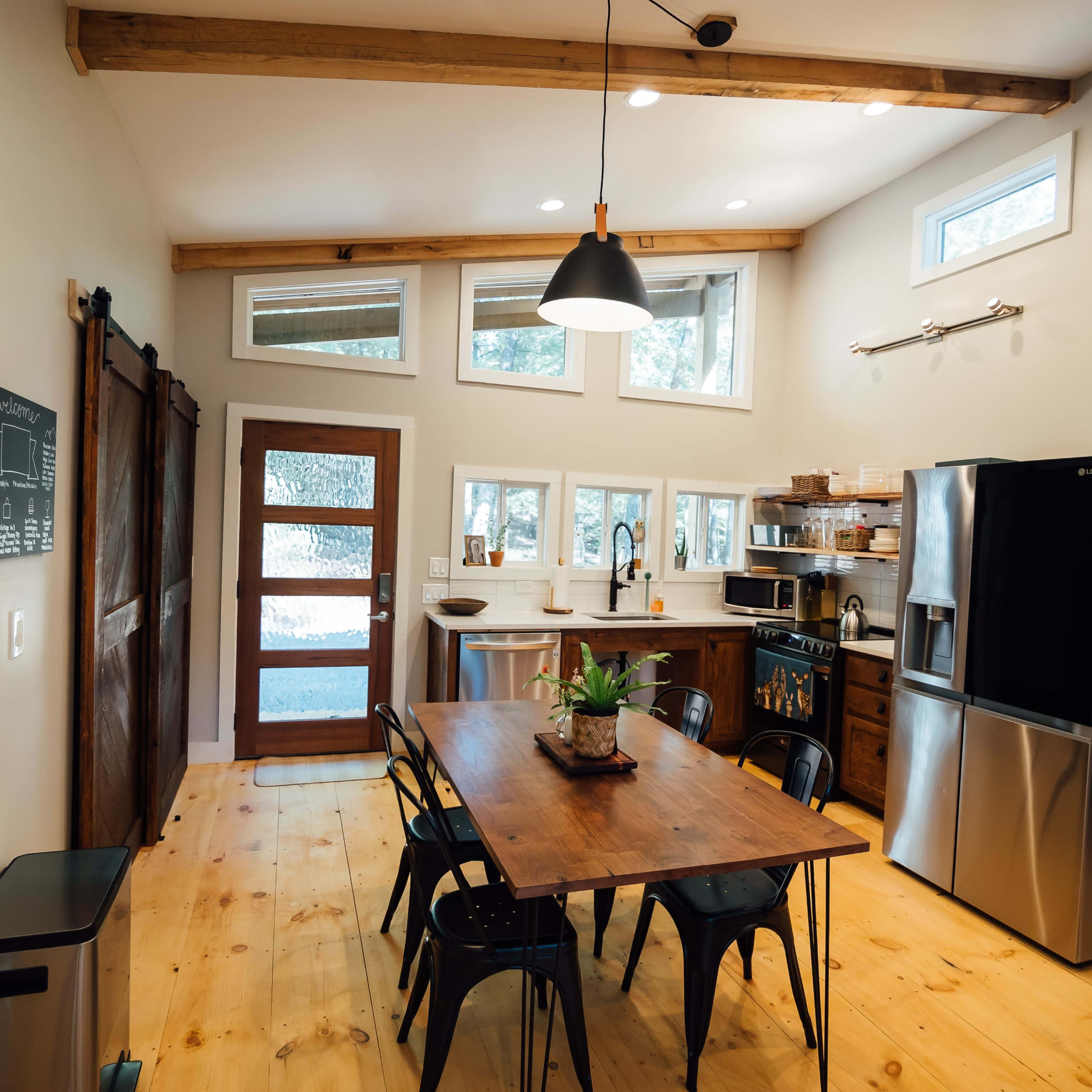 A modern kitchen with a wooden dining table, stainless steel refrigerator, and a large window allowing natural light to enter.