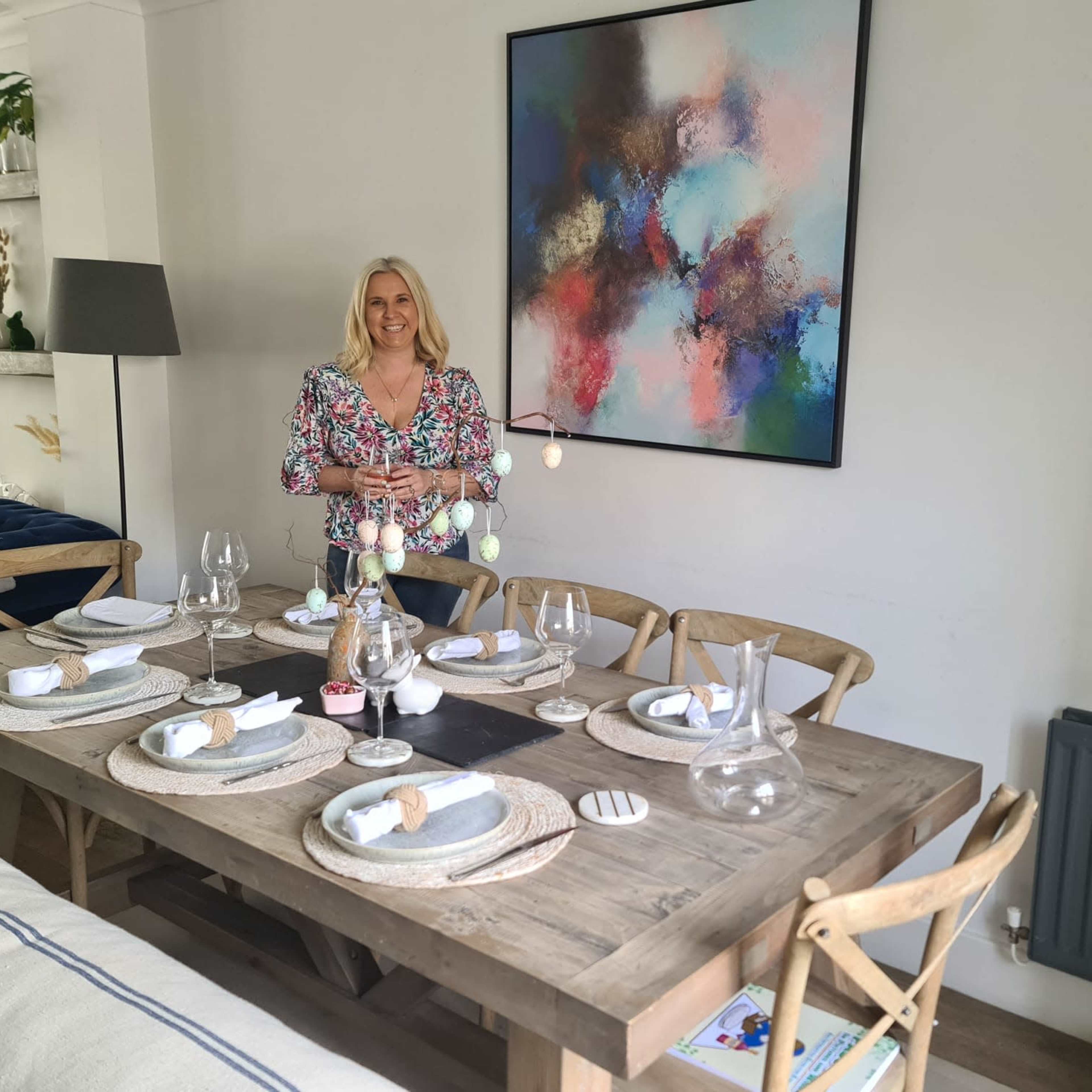A woman stands next to a dining table set with plates and glasses, with a colorful abstract painting on the wall behind her.