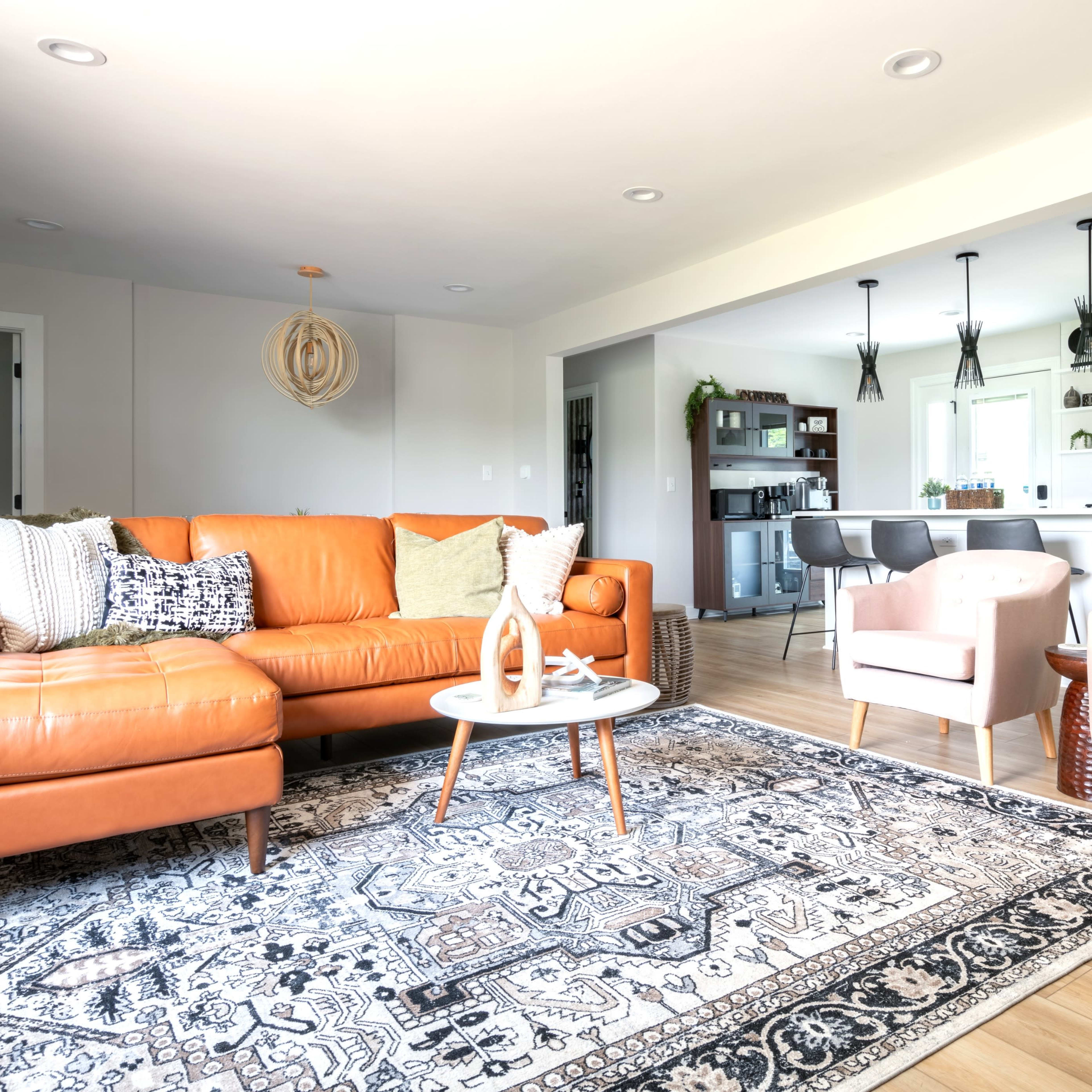 A modern living room features an orange leather sofa, two light pink armchairs, a decorative rug, and a view of a kitchen area in the background.