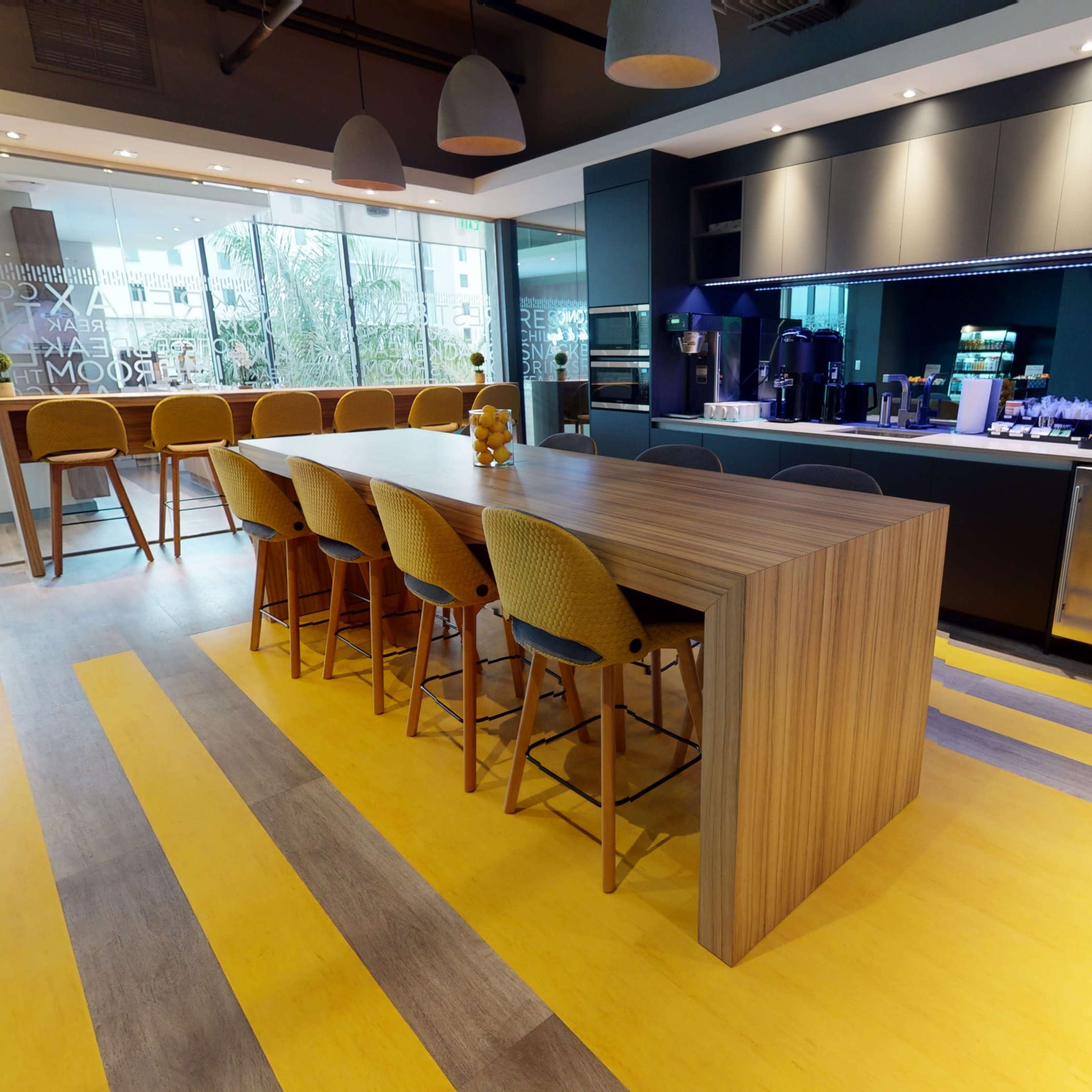 A modern kitchen area features a long wooden island with bar stools, a coffee station, and large windows allowing natural light to enter.