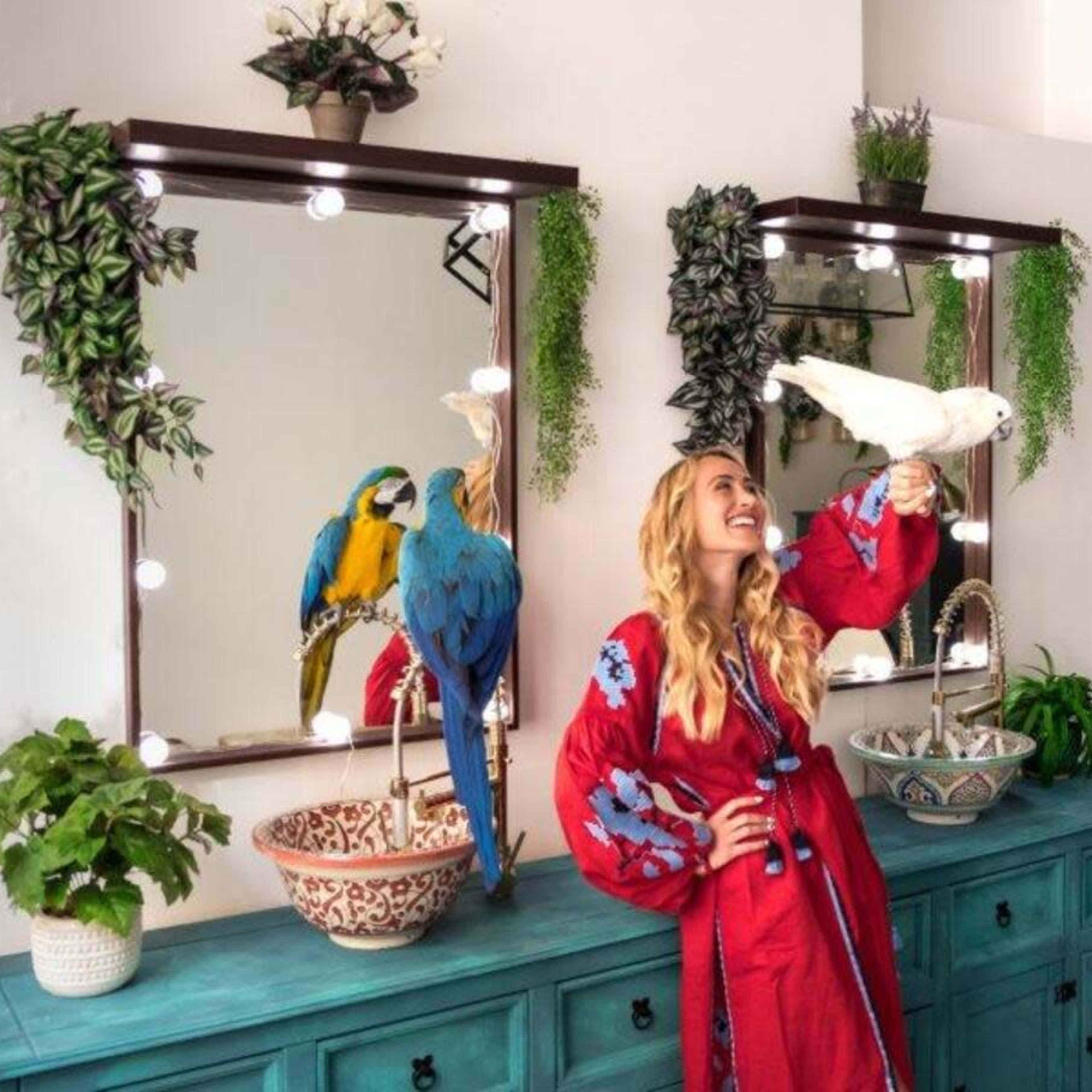 A woman in a red robe stands in front of a decorated vanity with two colorful birds perched on her hand and nearby mirrors.
