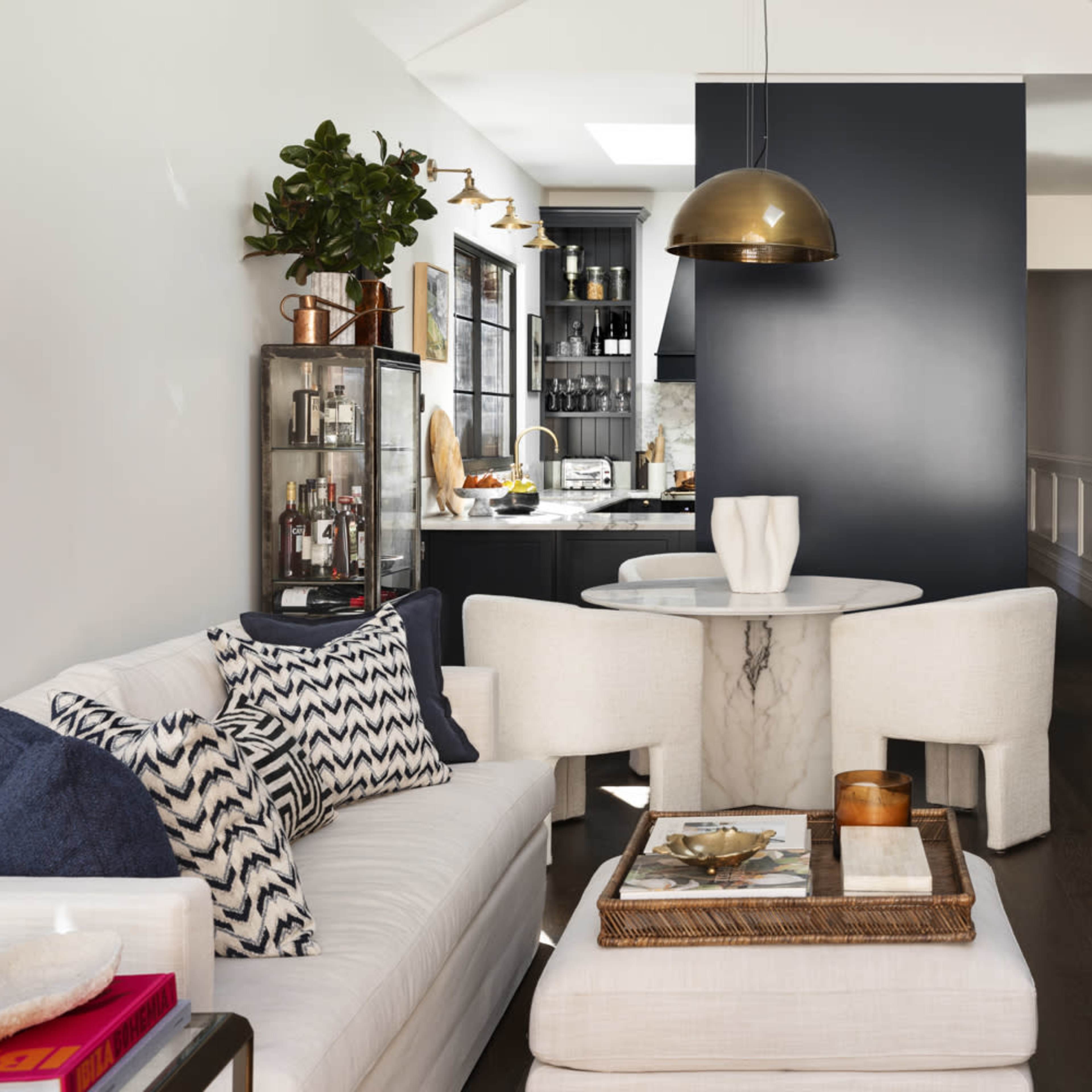 A modern living space features a white sofa with patterned cushions, a round marble table, and a dark accent wall leading to a kitchen area.