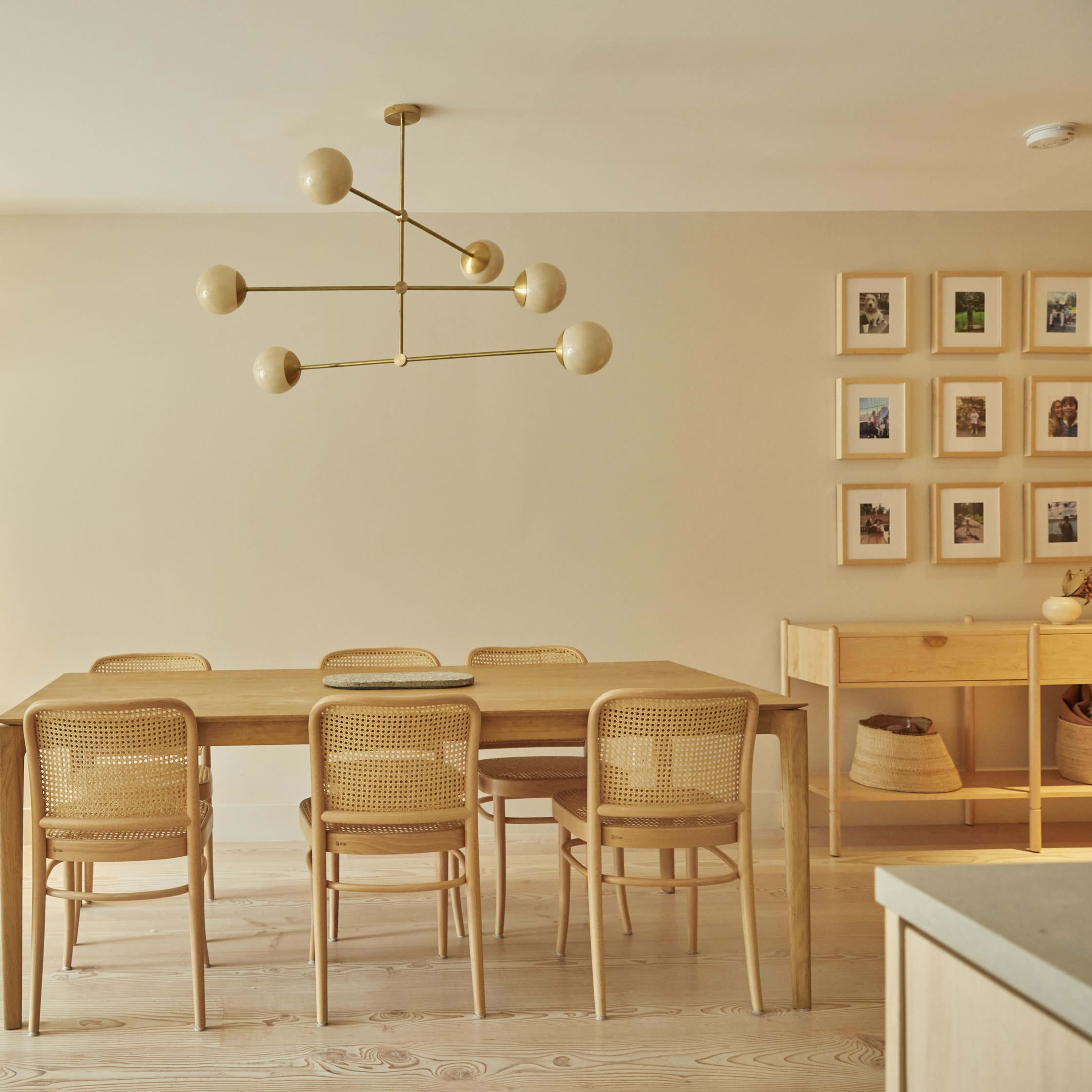 A modern dining area features a wooden table surrounded by six chairs, a light fixture with multiple bulbs overhead, and shelves with decorative items and framed photos on the walls.