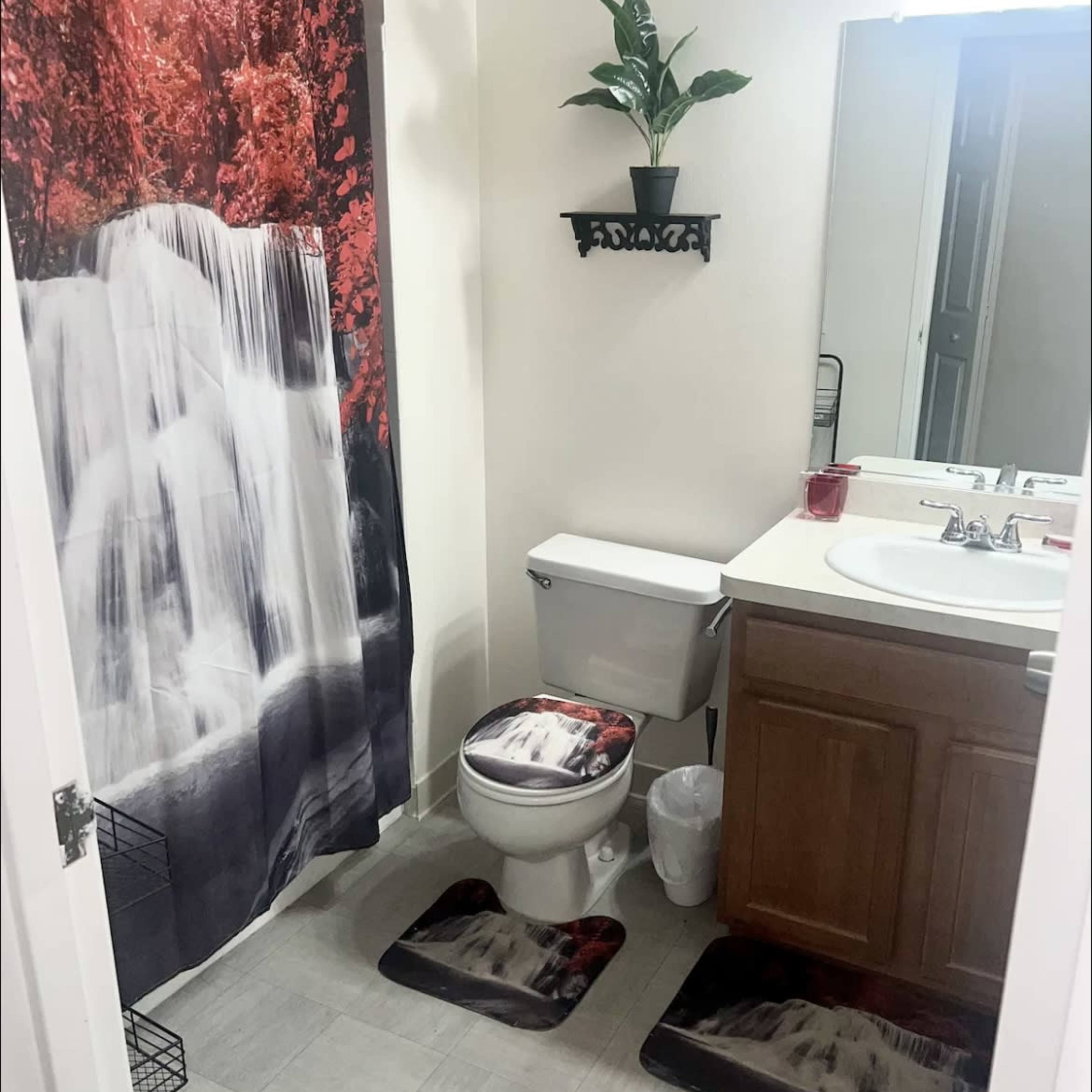 A bathroom with a waterfall-themed shower curtain, a toilet, a sink with a wooden cabinet, and matching floor mats.