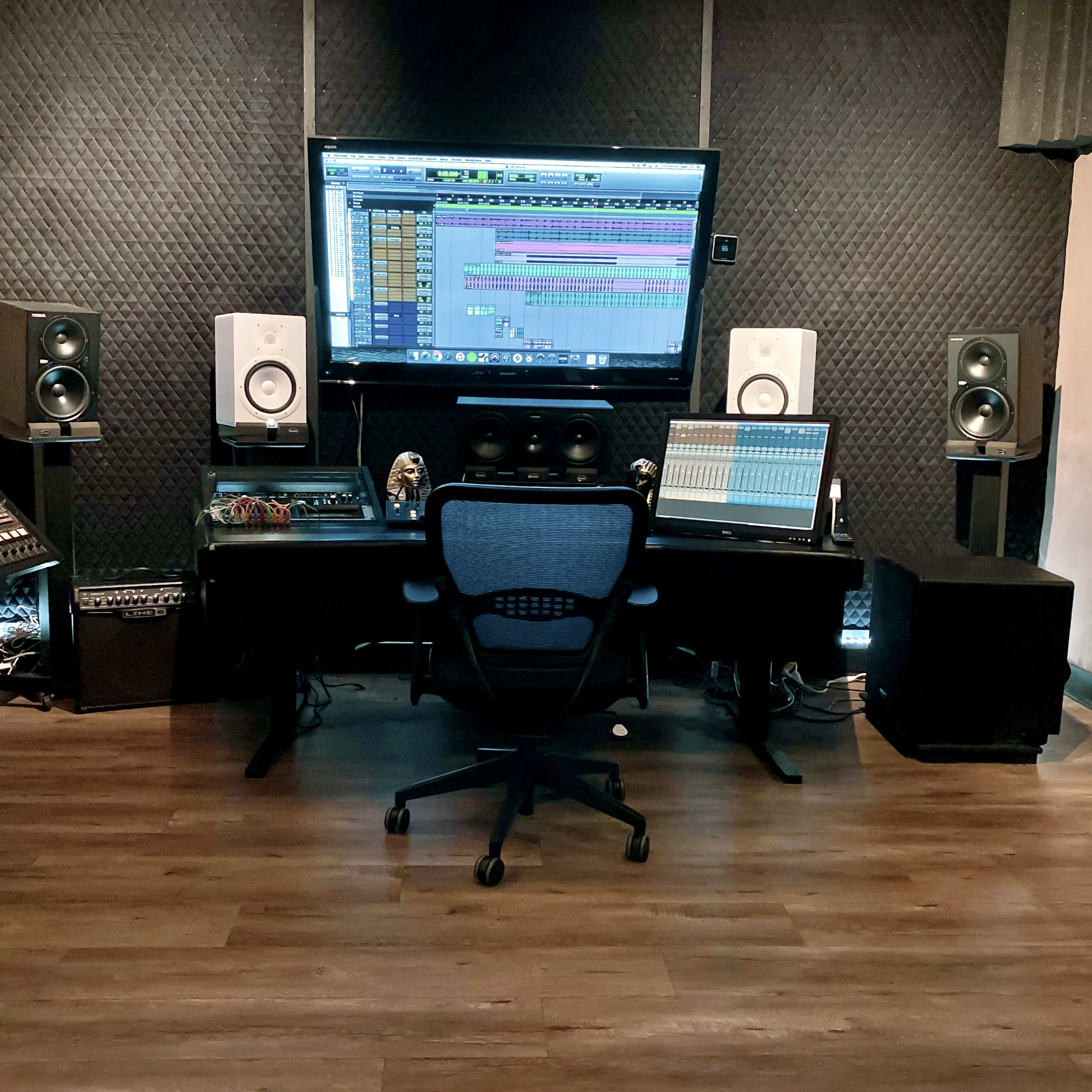 A music studio featuring a computer monitor displaying audio software, a digital audio workstation, studio speakers, and various musical equipment arranged around a central swivel chair.
