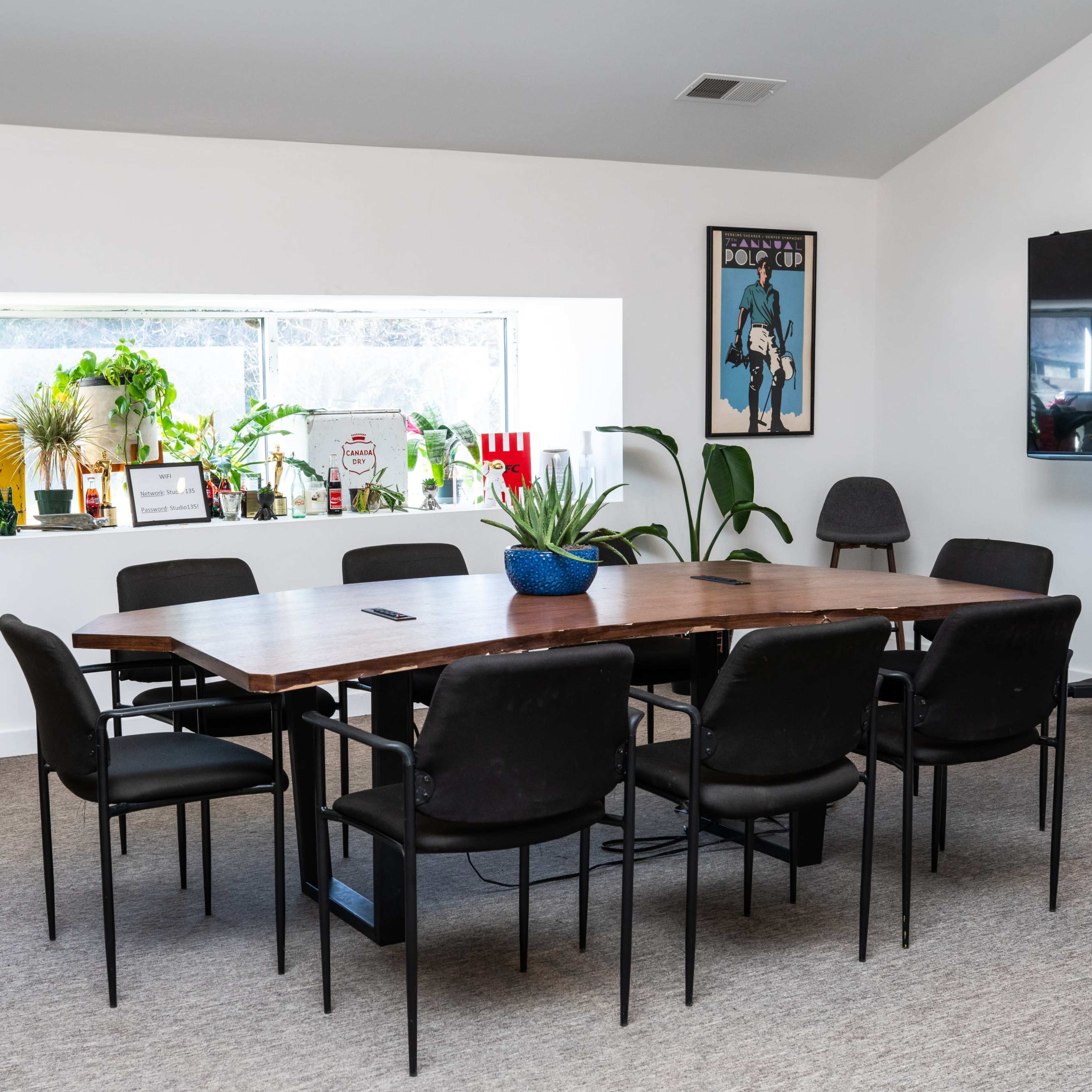 A long wooden table surrounded by black chairs is positioned in a well-lit meeting room with potted plants and decorative items on the windowsill.