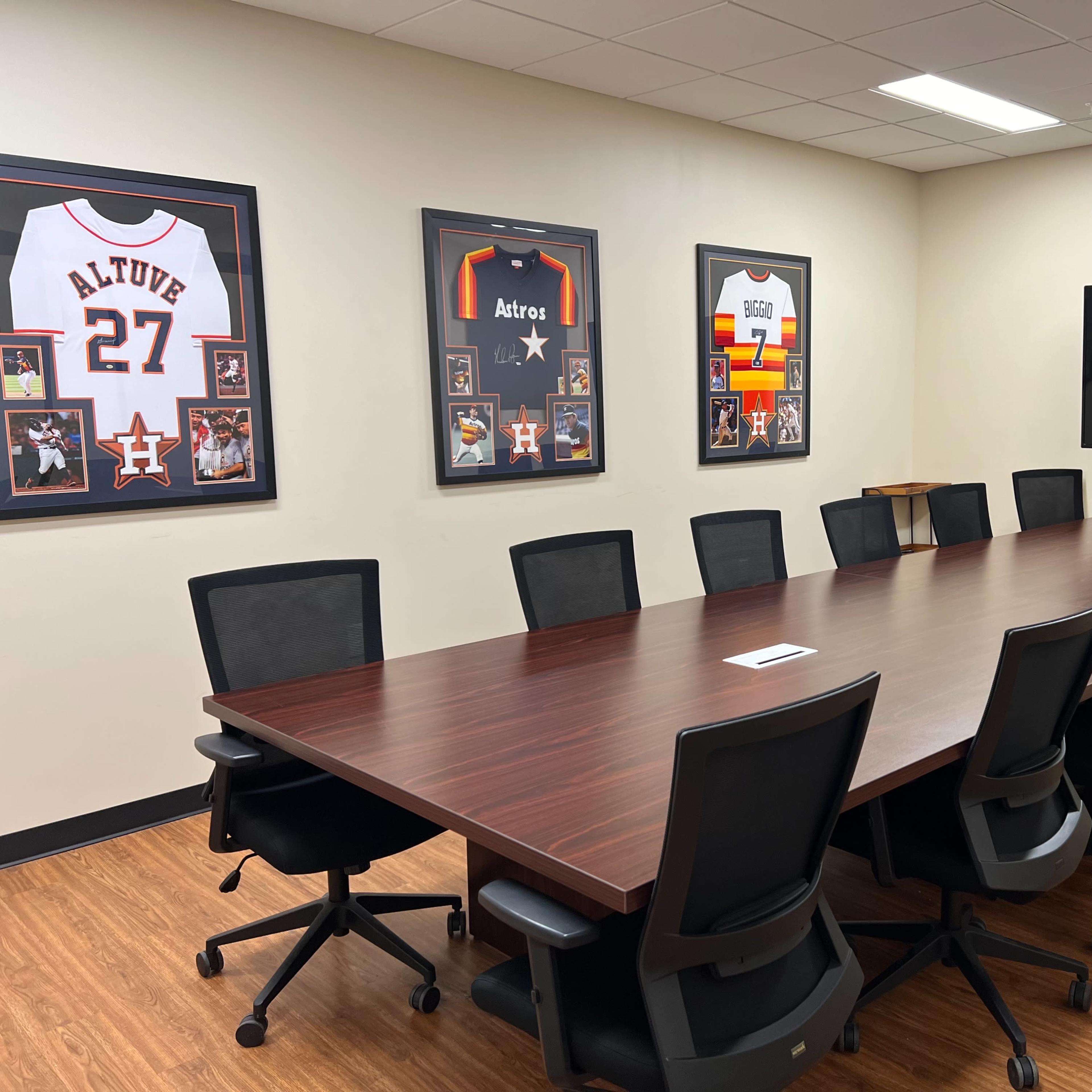 A conference room with a long wooden table surrounded by black office chairs, adorned with framed jerseys and a large television on the wall.