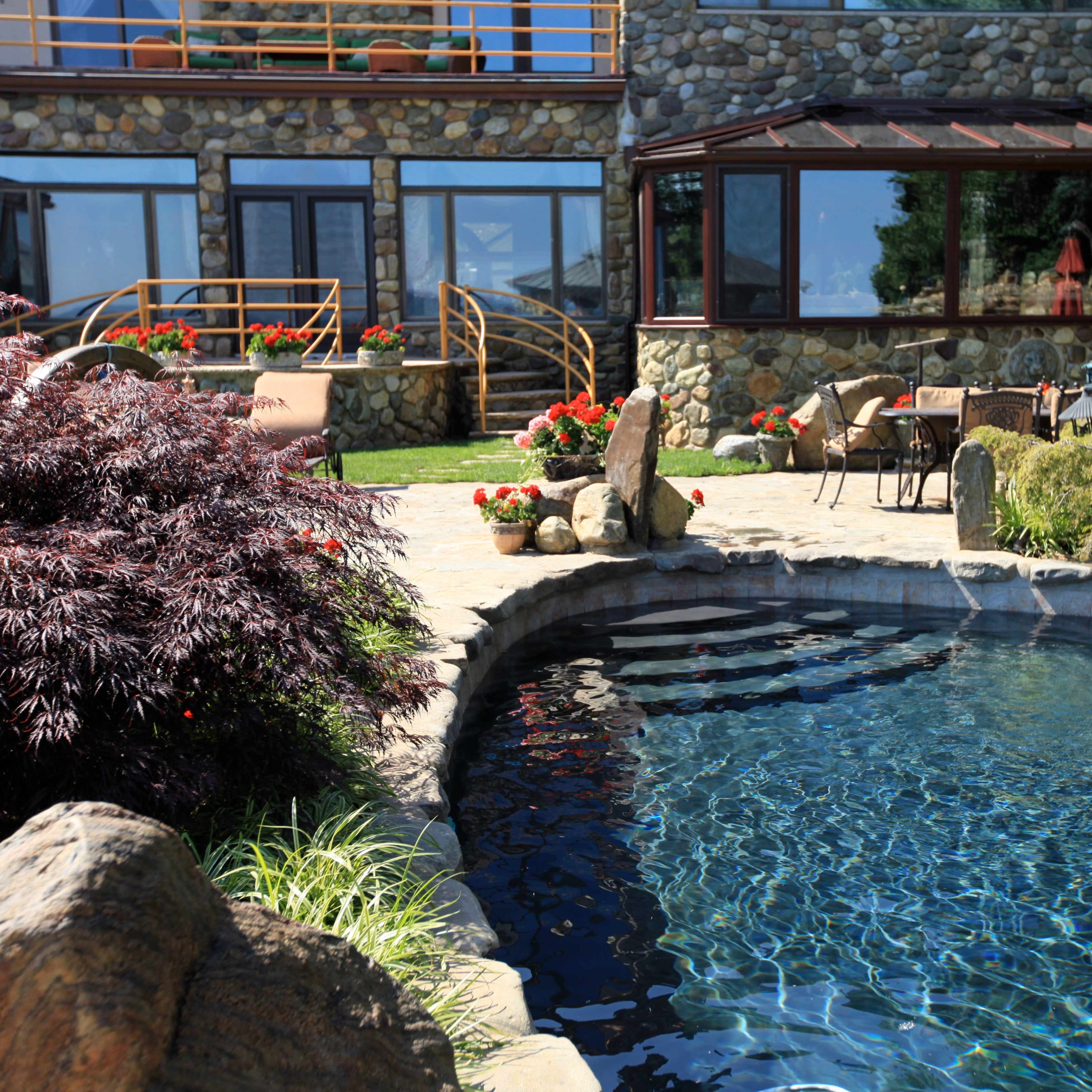 A stone patio with a swimming pool is surrounded by landscaped gardens and a rustic building.