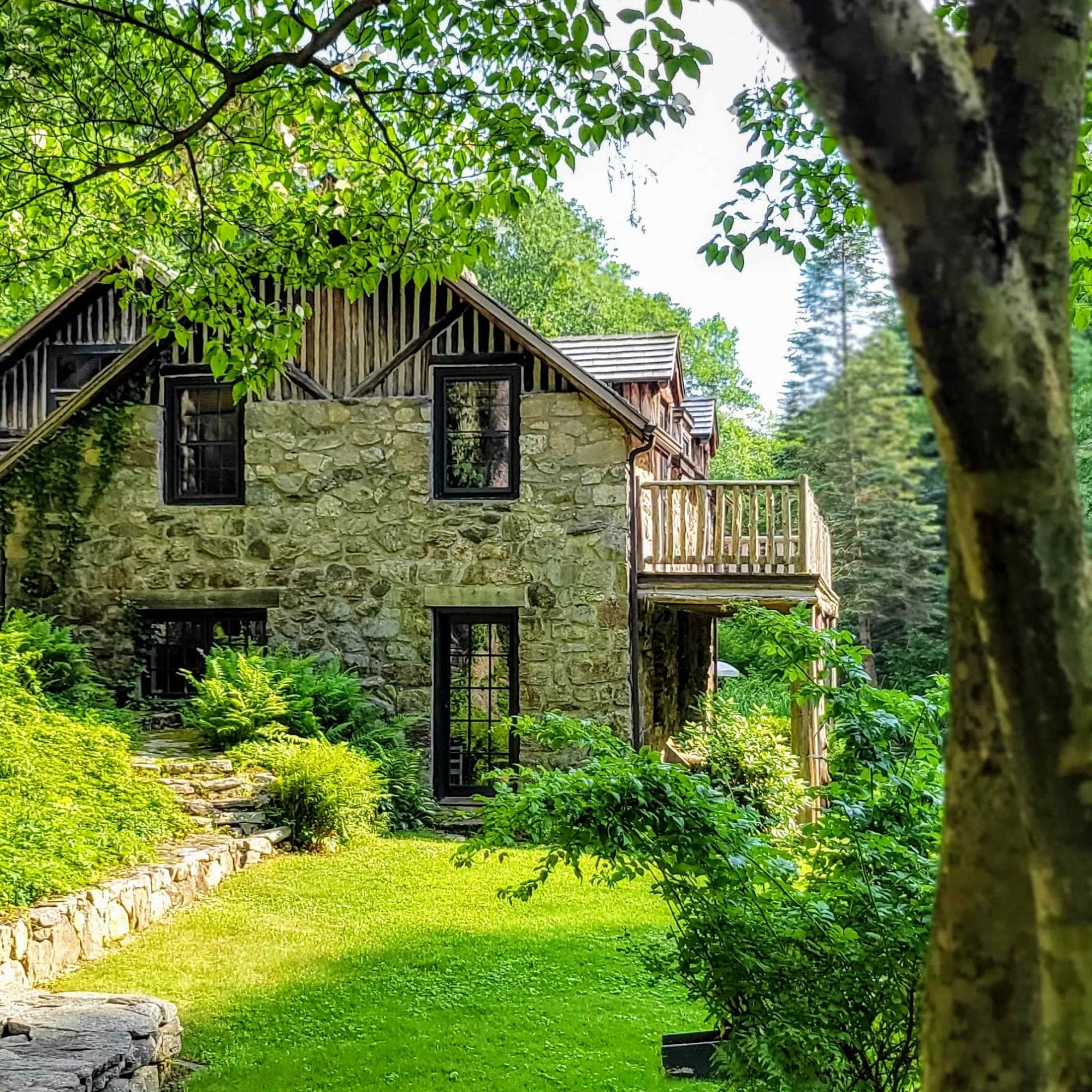 A stone house with large windows and a balcony is nestled among greenery in a landscaped yard.
