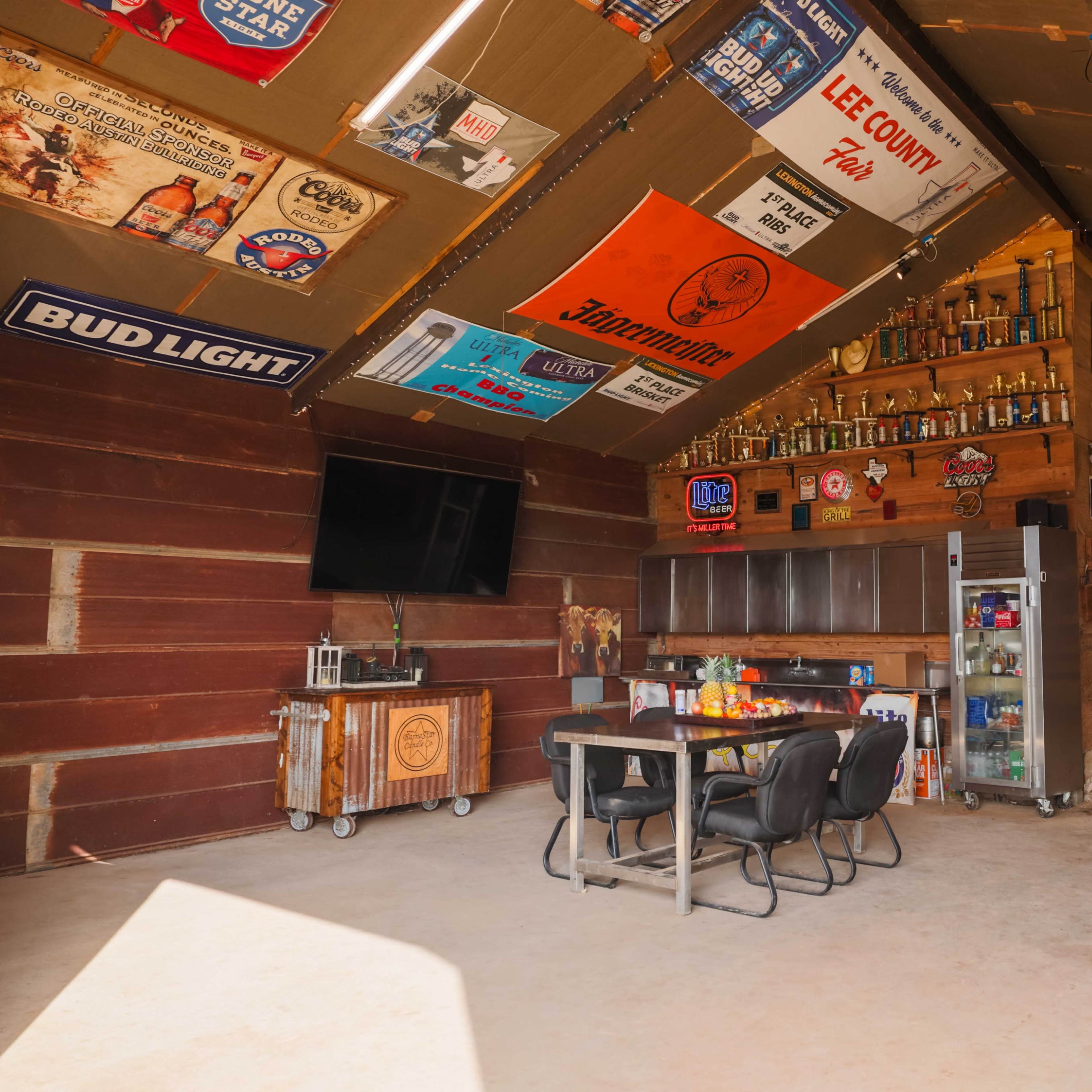 An interior of a rustic gathering space with wood-paneled walls, a table and chairs, a television mounted to one side, and various trophies and signs displayed overhead.