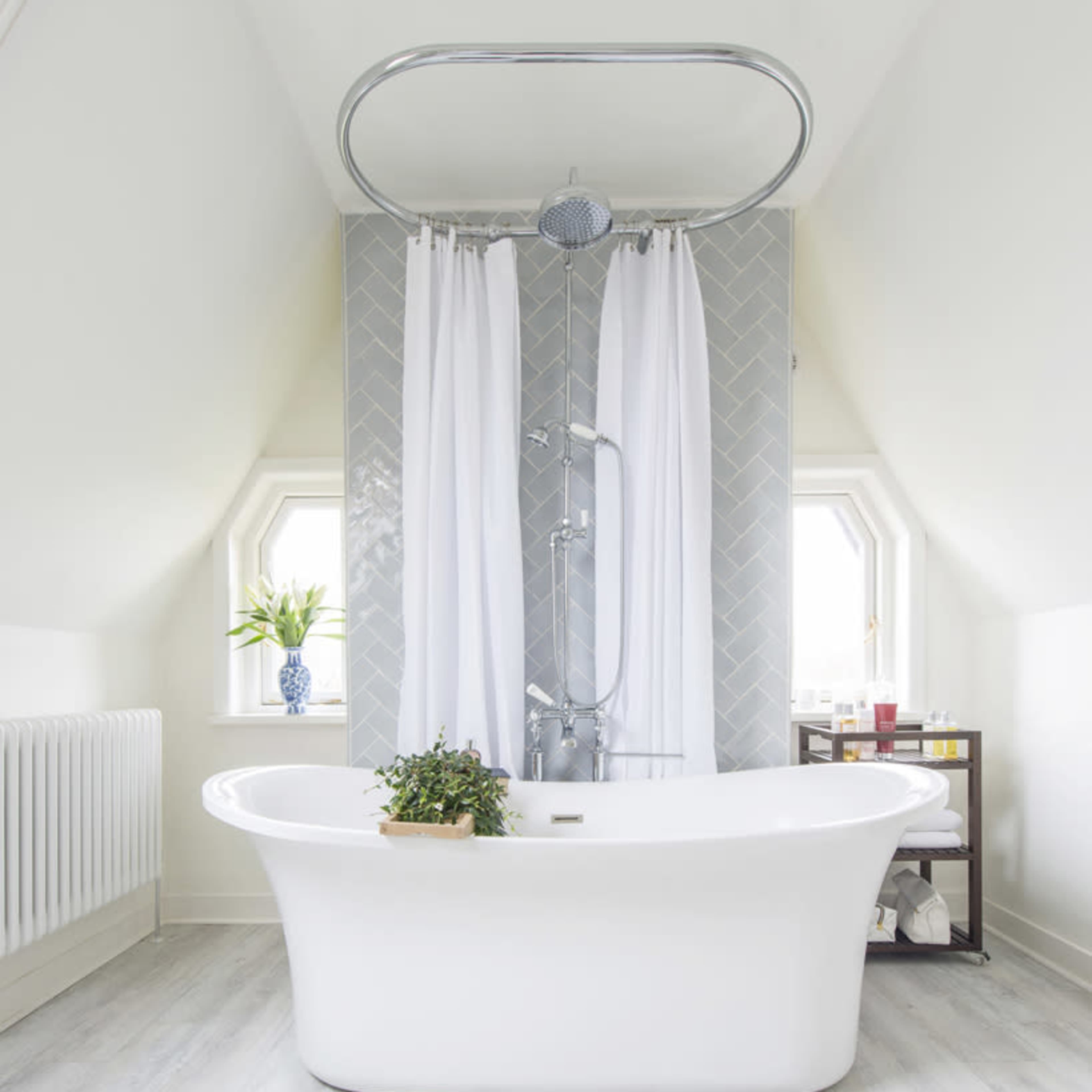 A freestanding white bathtub is positioned in a bright, airy bathroom with white curtains, a tiled shower area, and a small plant on the ledge.