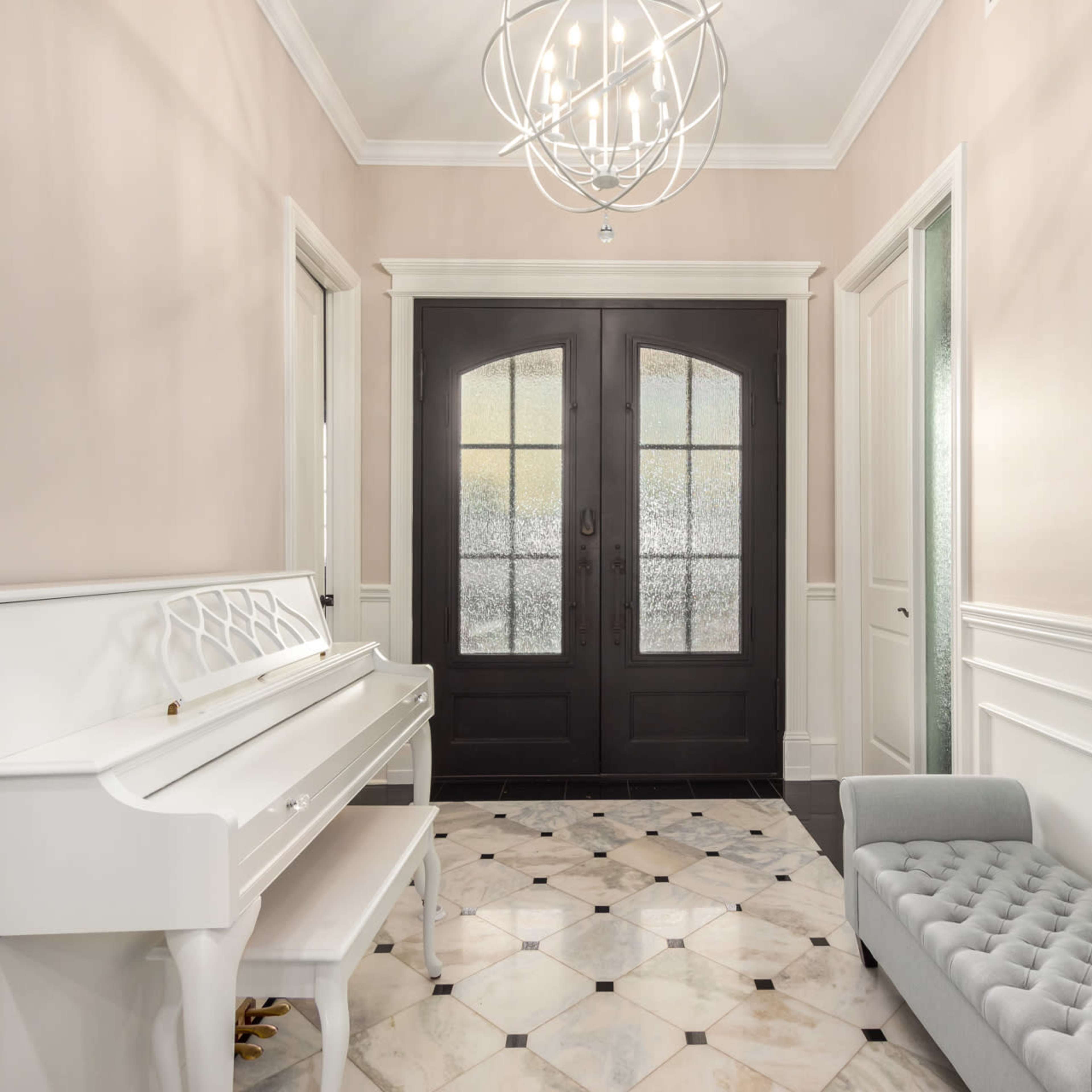 A hallway with a white piano, a gray tufted bench, and black double doors, featuring marble flooring and a modern chandelier.