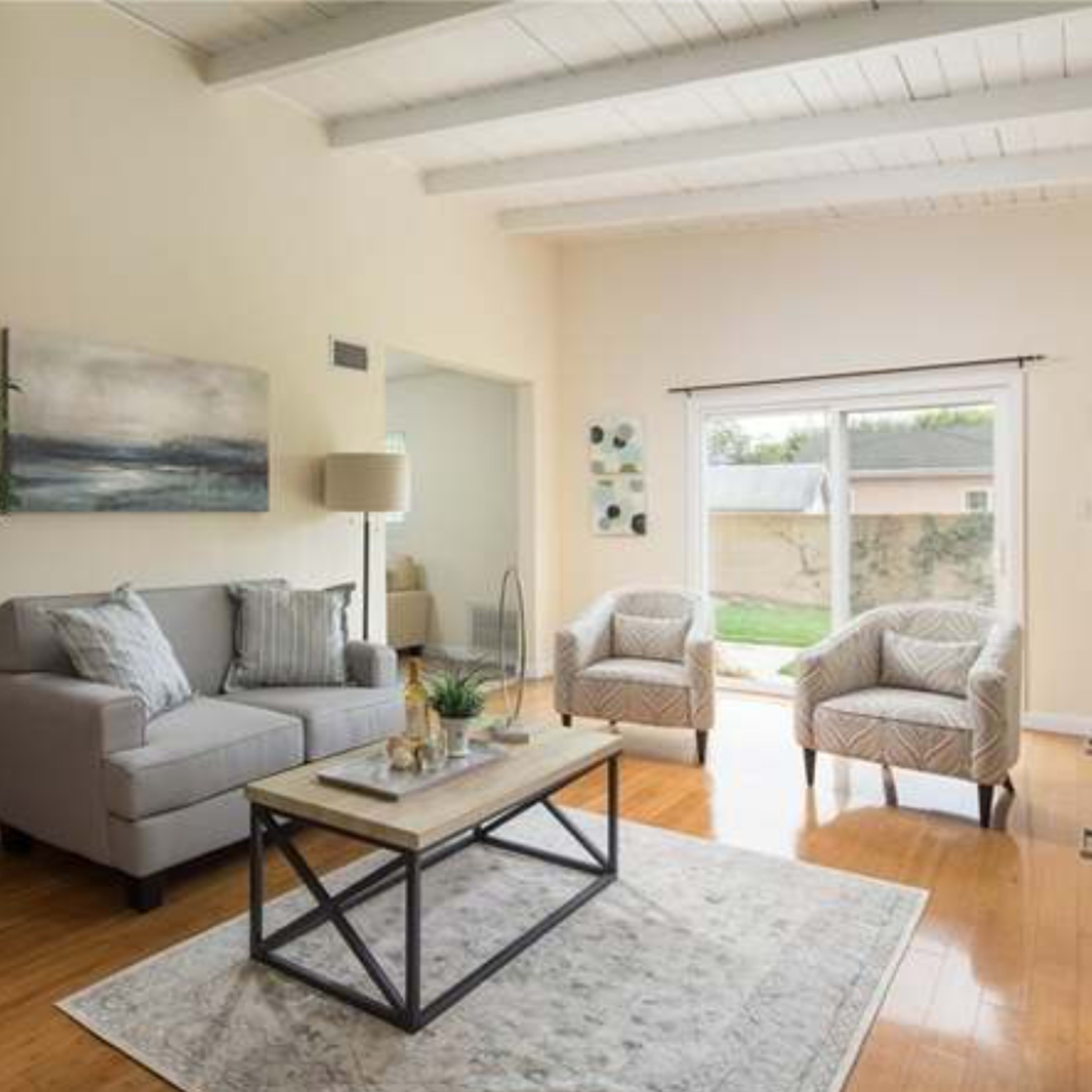 A bright living room features a gray sofa, two patterned chairs, a coffee table, and a view of the backyard through sliding glass doors.