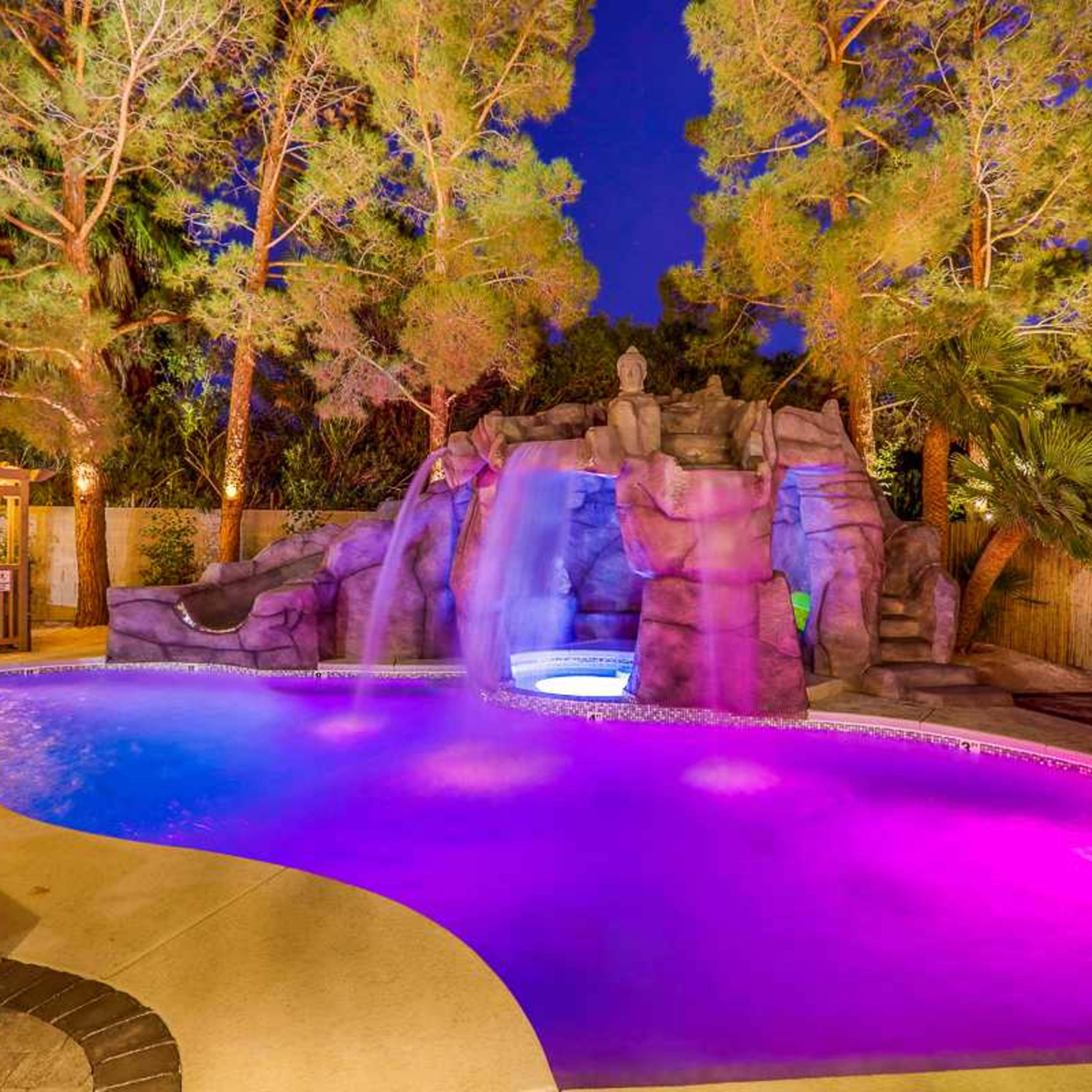 A backyard pool features a waterfall cascading over rocky terrain, surrounded by trees and illuminated with vibrant purple lights.