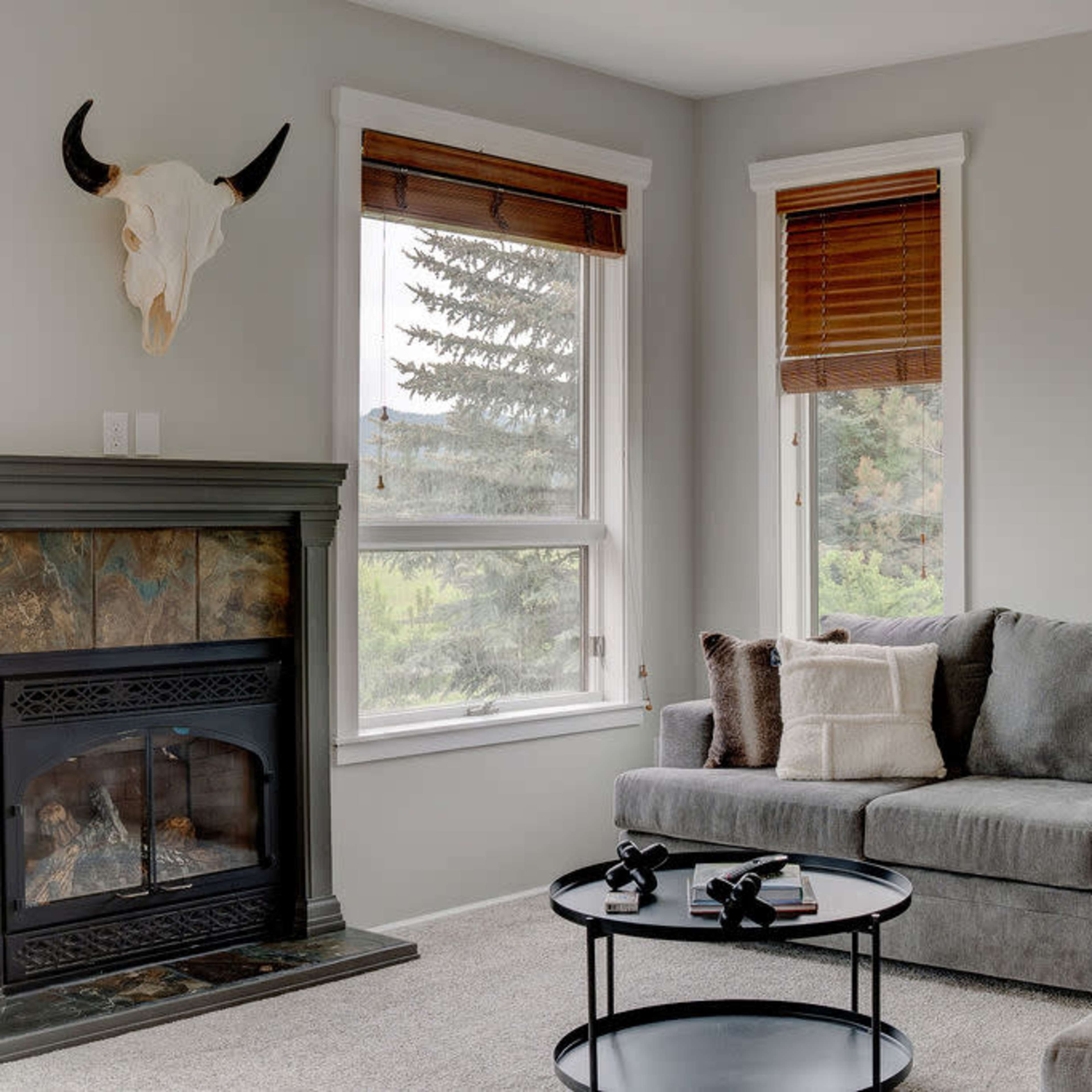 The living room features a gray sofa, a black coffee table with decorative items, a fireplace, and large windows with wooden blinds.