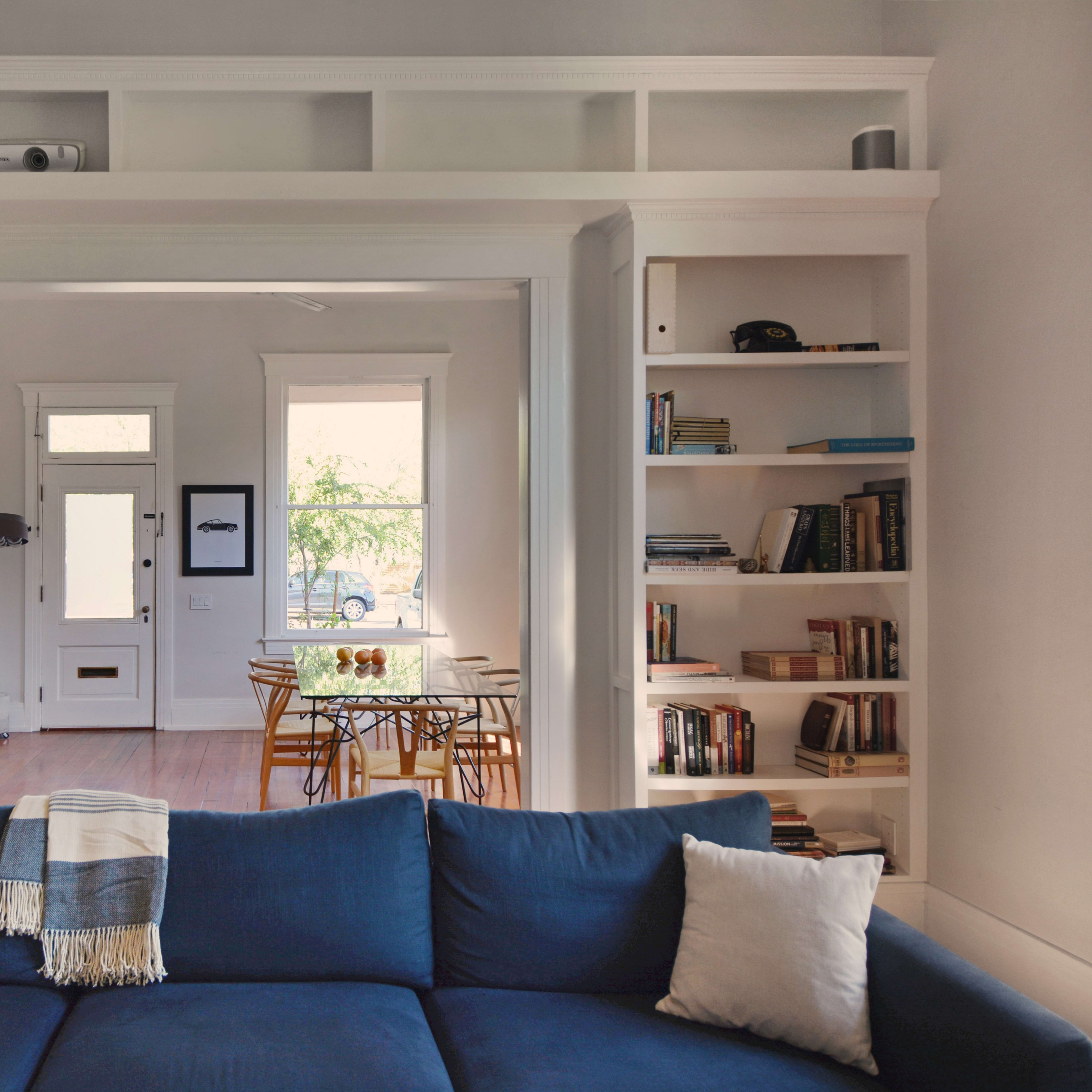 A living room features a large blue sofa facing built-in bookshelves, with a doorway leading to a dining area visible in the background.