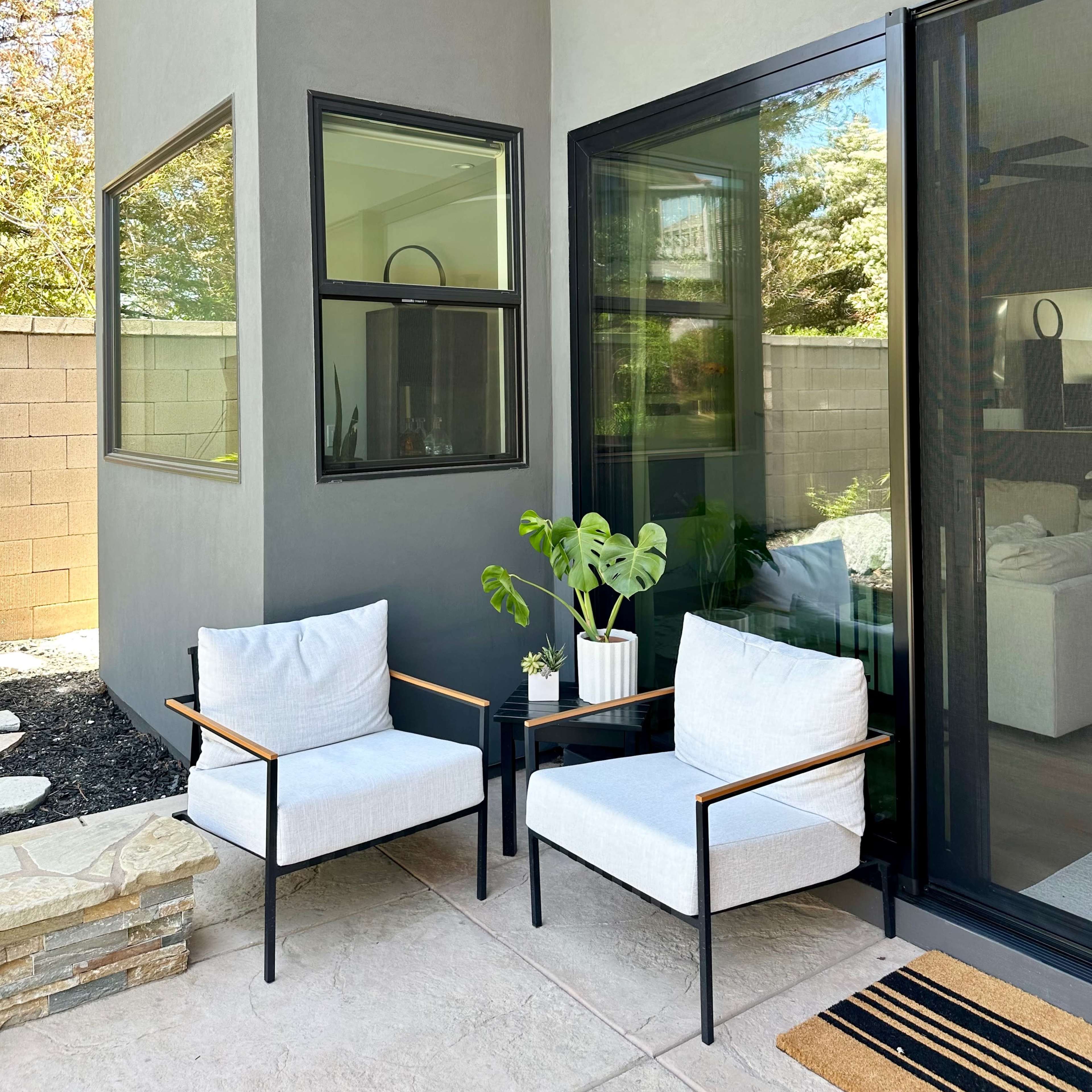 Two modern chairs and a small table are positioned outside a contemporary home next to large windows.