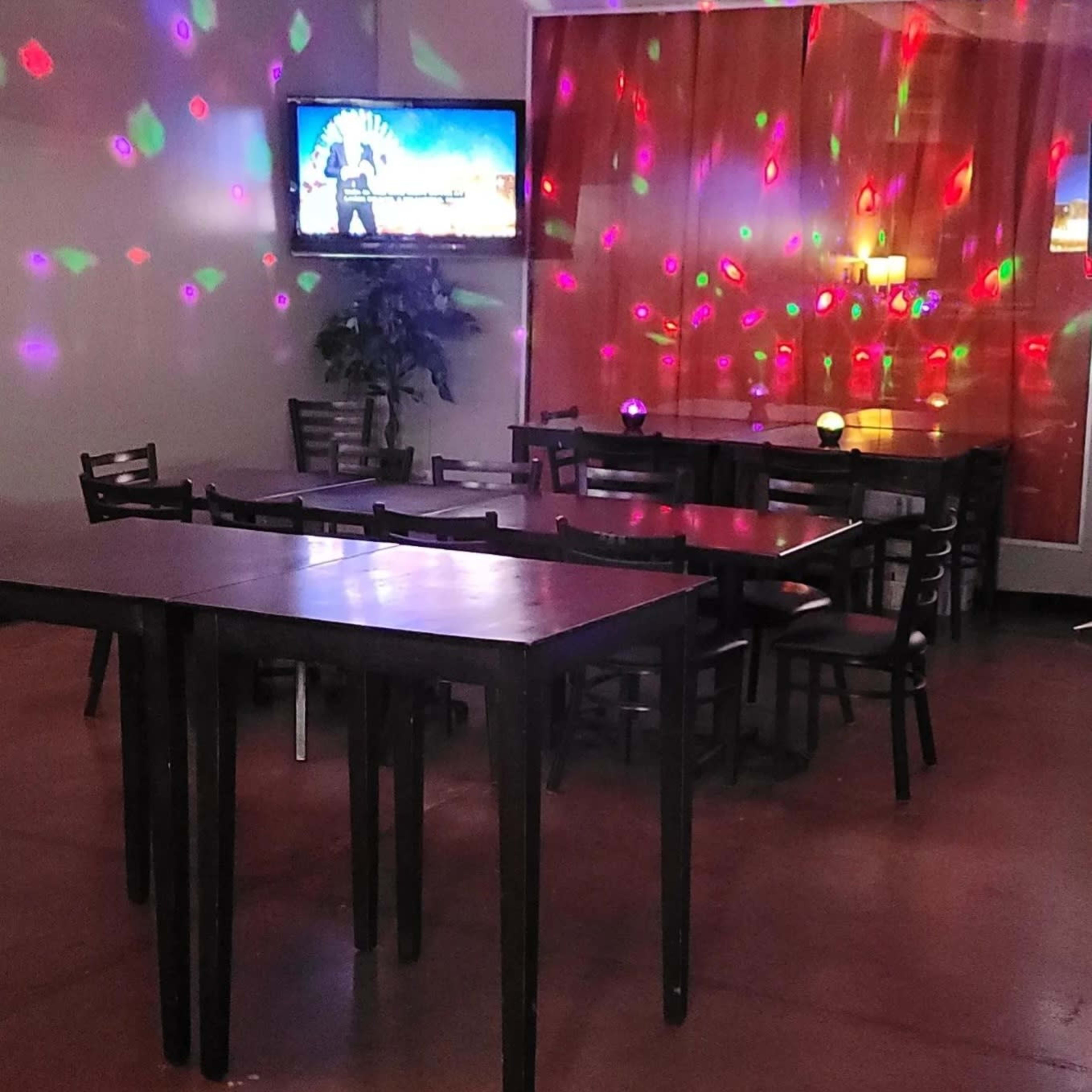 A dimly lit interior of a venue with multiple tables, a TV displaying a performance, and colorful lights reflecting on the walls.
