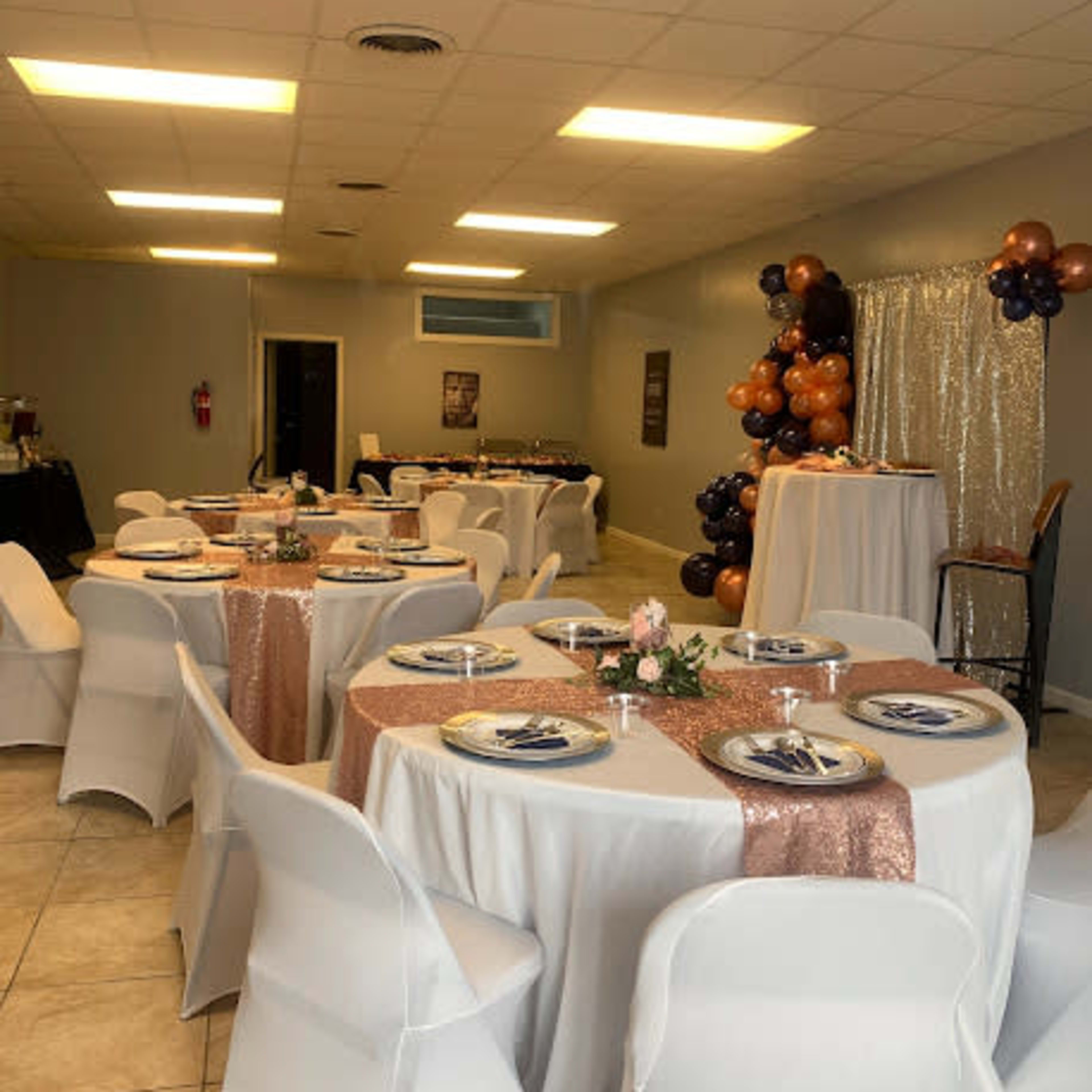 A banquet hall is set up with several tables covered in white tablecloths and decorated with rose gold accents, surrounded by chairs and a backdrop featuring balloons.