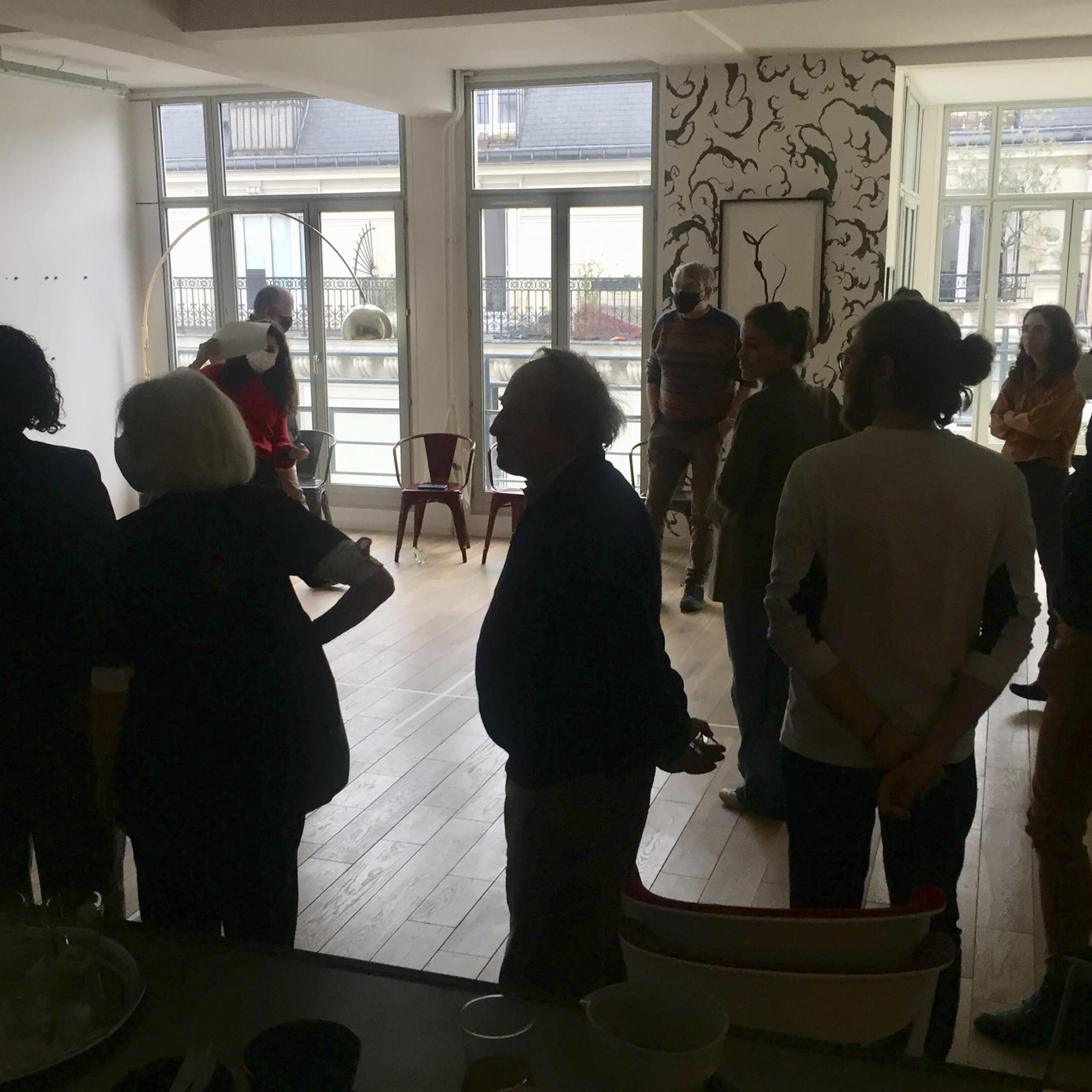 A group of people stands in a well-lit room with large windows and a decorative wall, engaging in conversation.