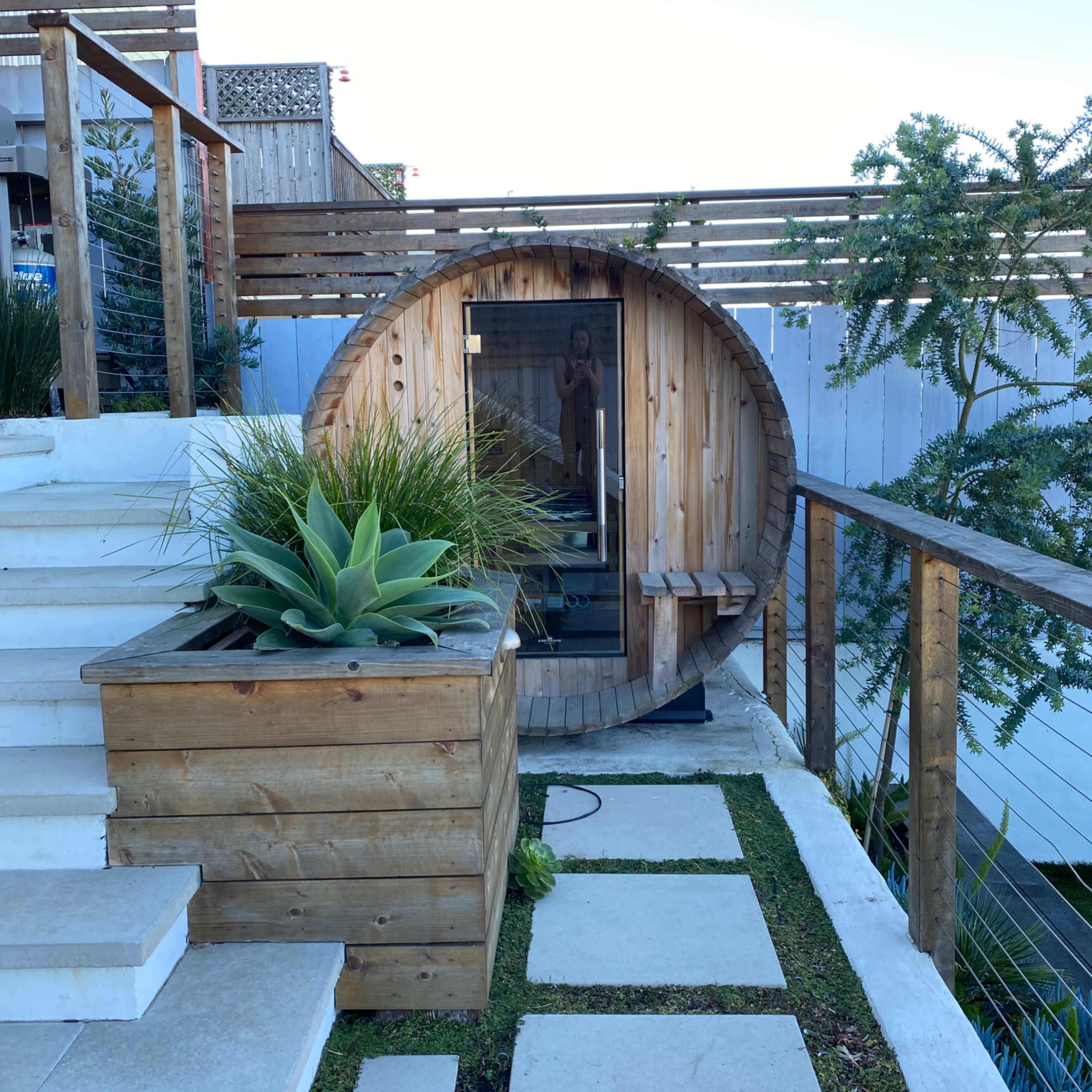 A wooden barrel sauna is positioned next to stone steps and a planter featuring tall grass and succulents.