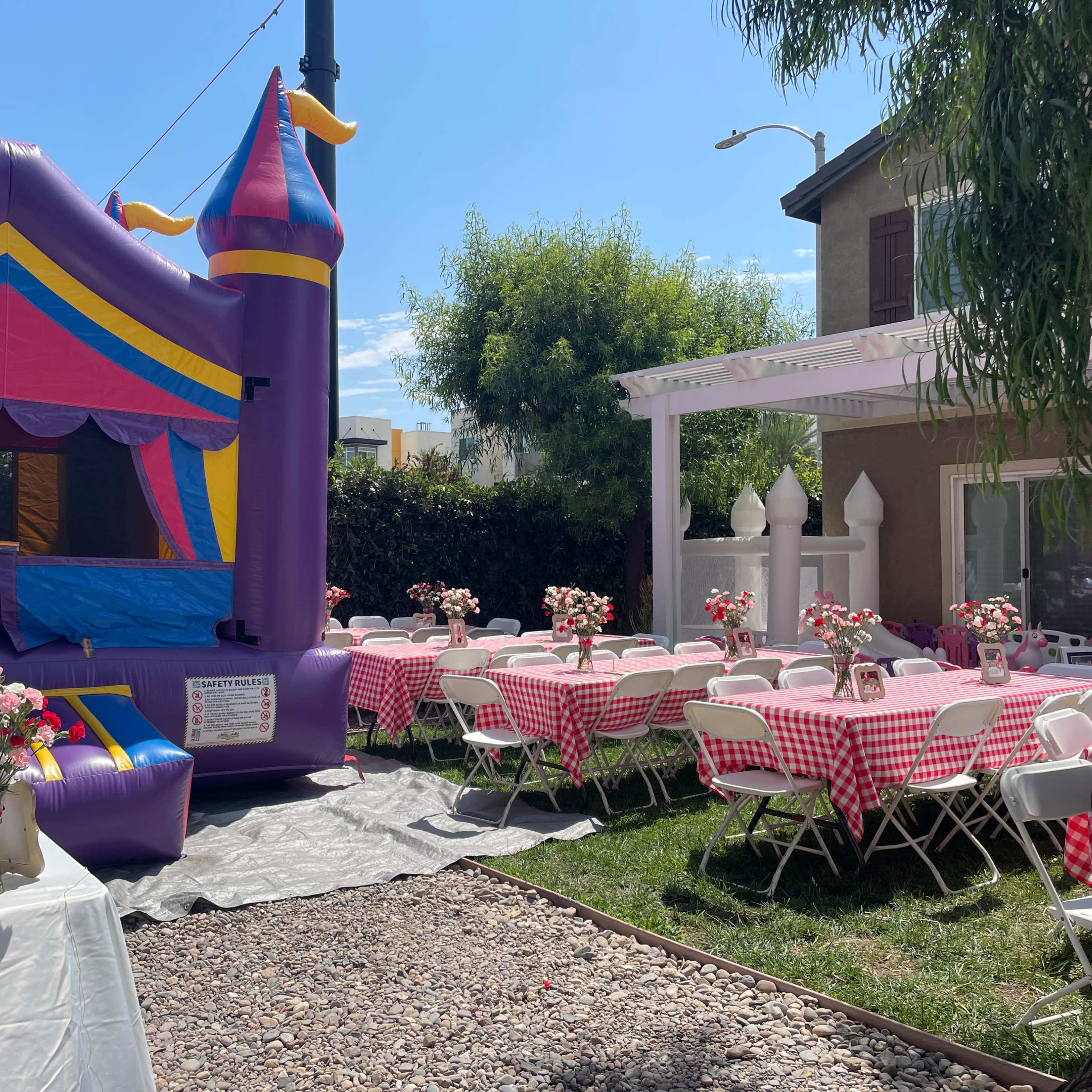 A backyard party setup features a colorful bounce house and tables dressed in checkered tablecloths surrounded by decorations and flowers.