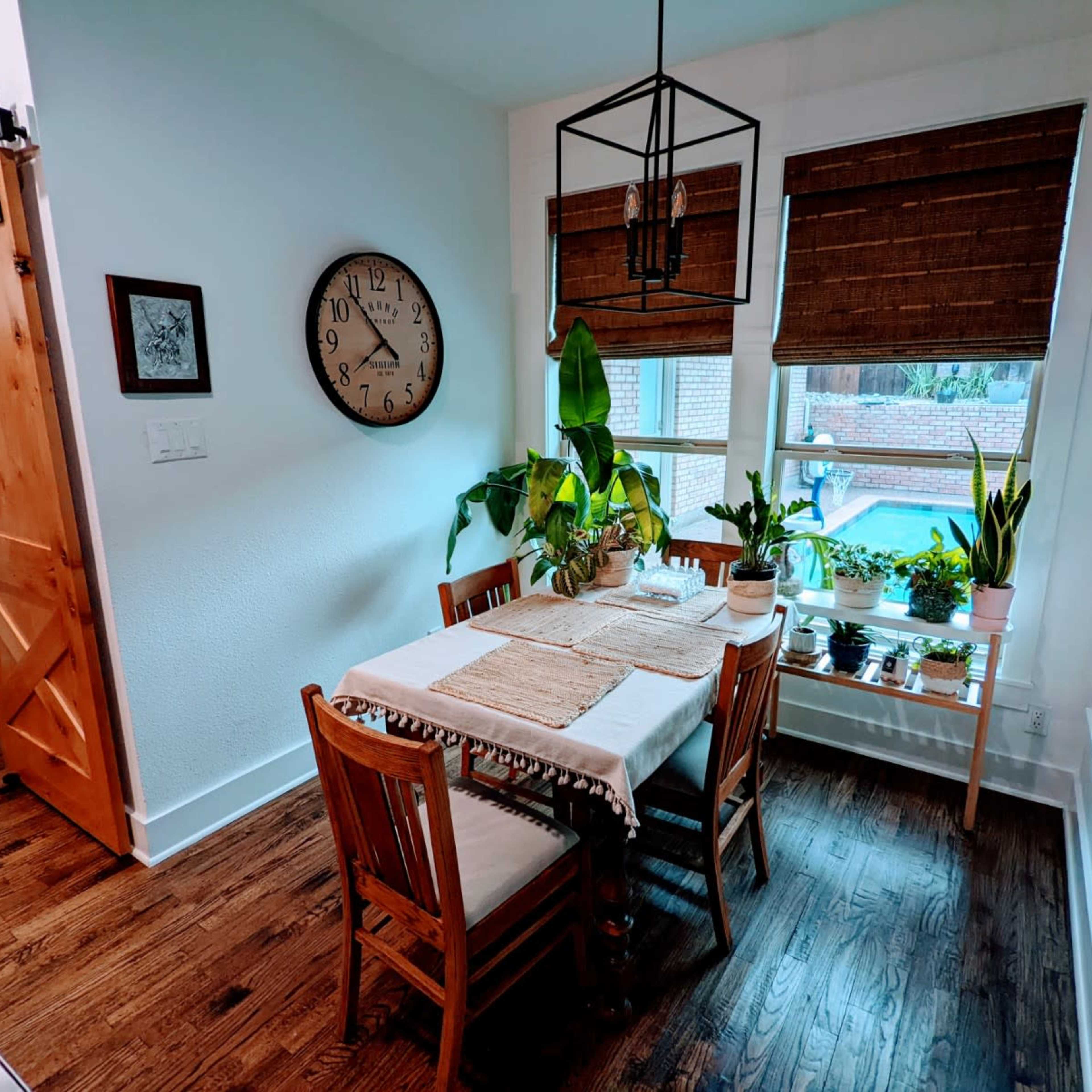A small dining area features a wooden table with four chairs, surrounded by potted plants and equipped with a large wall clock.