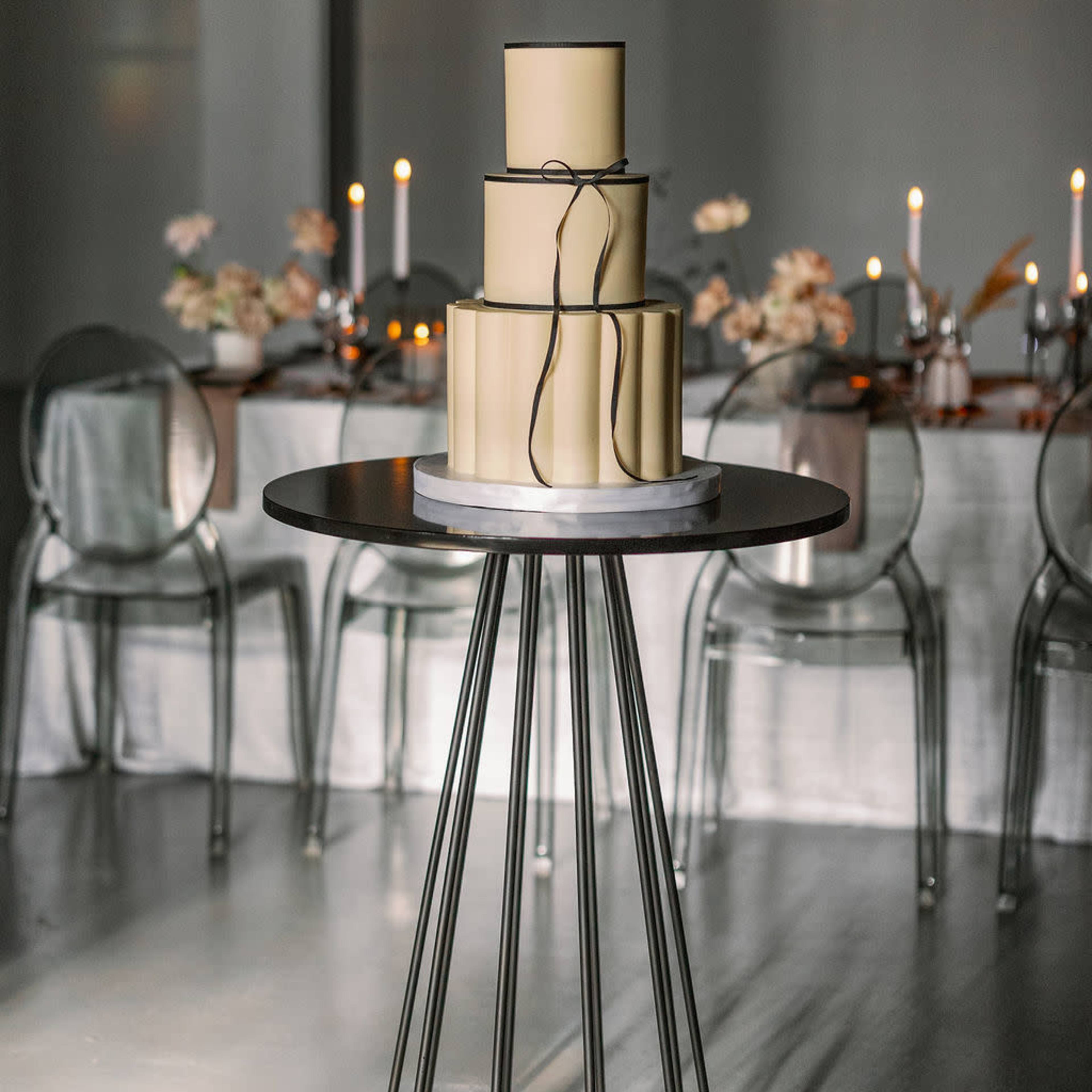 A three-tiered cake is placed on a circular table in a dimly lit room with a dining setup in the background.