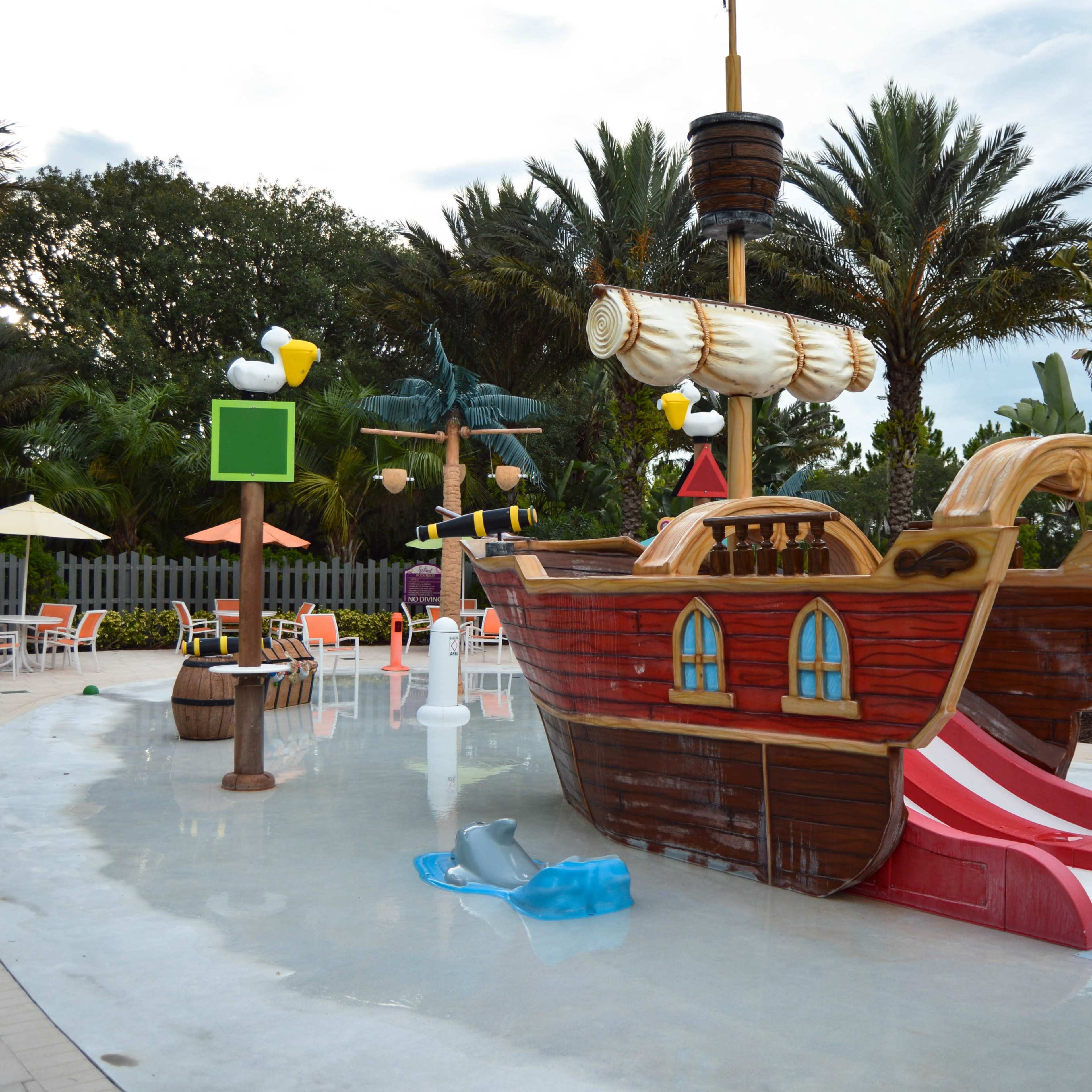 A large, colorful pirate ship water slide is situated in a splash pad area surrounded by palm trees and poolside lounge chairs.