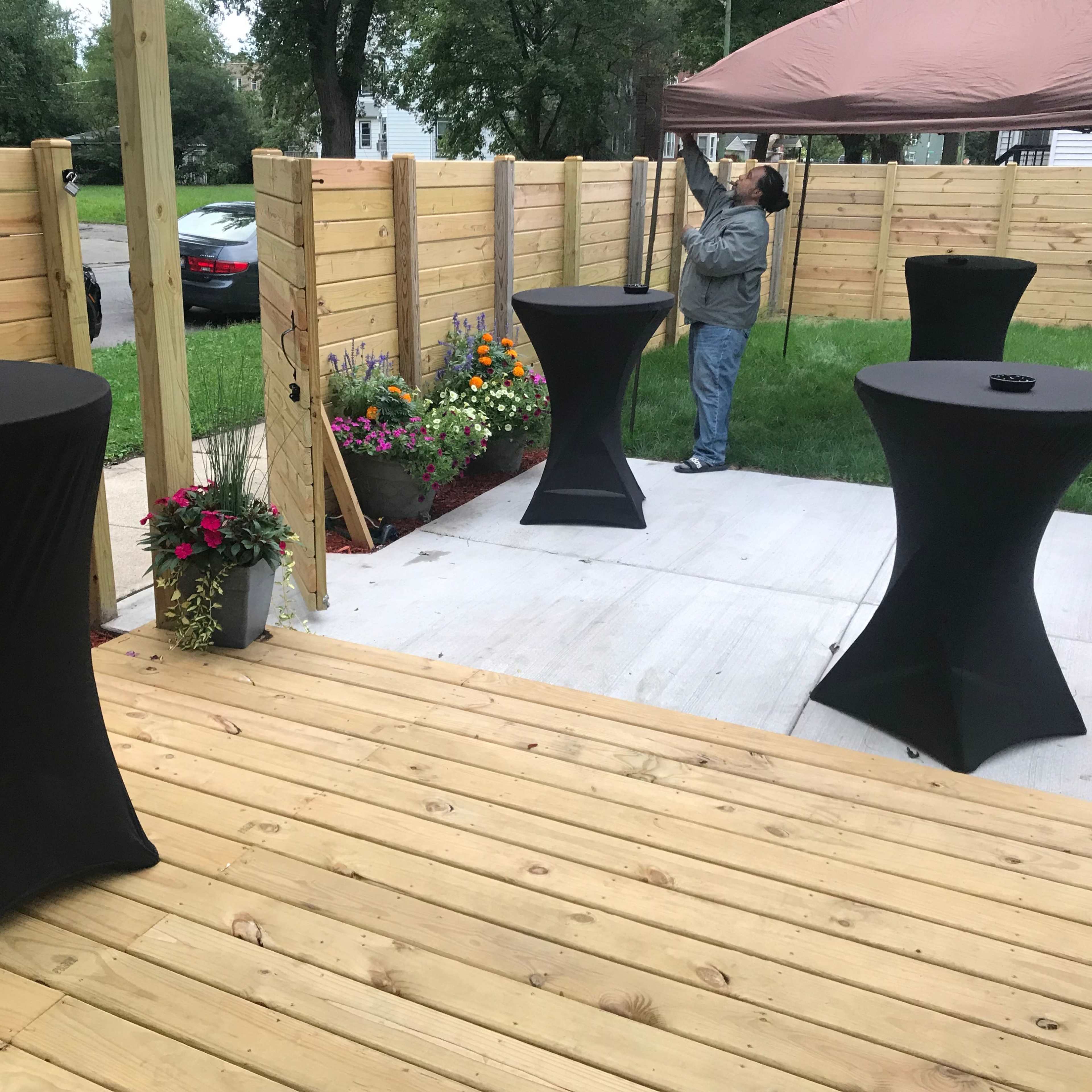 A patio area with three black cocktail tables and a man adjusting a tent in the background, surrounded by wooden fencing and flower beds.