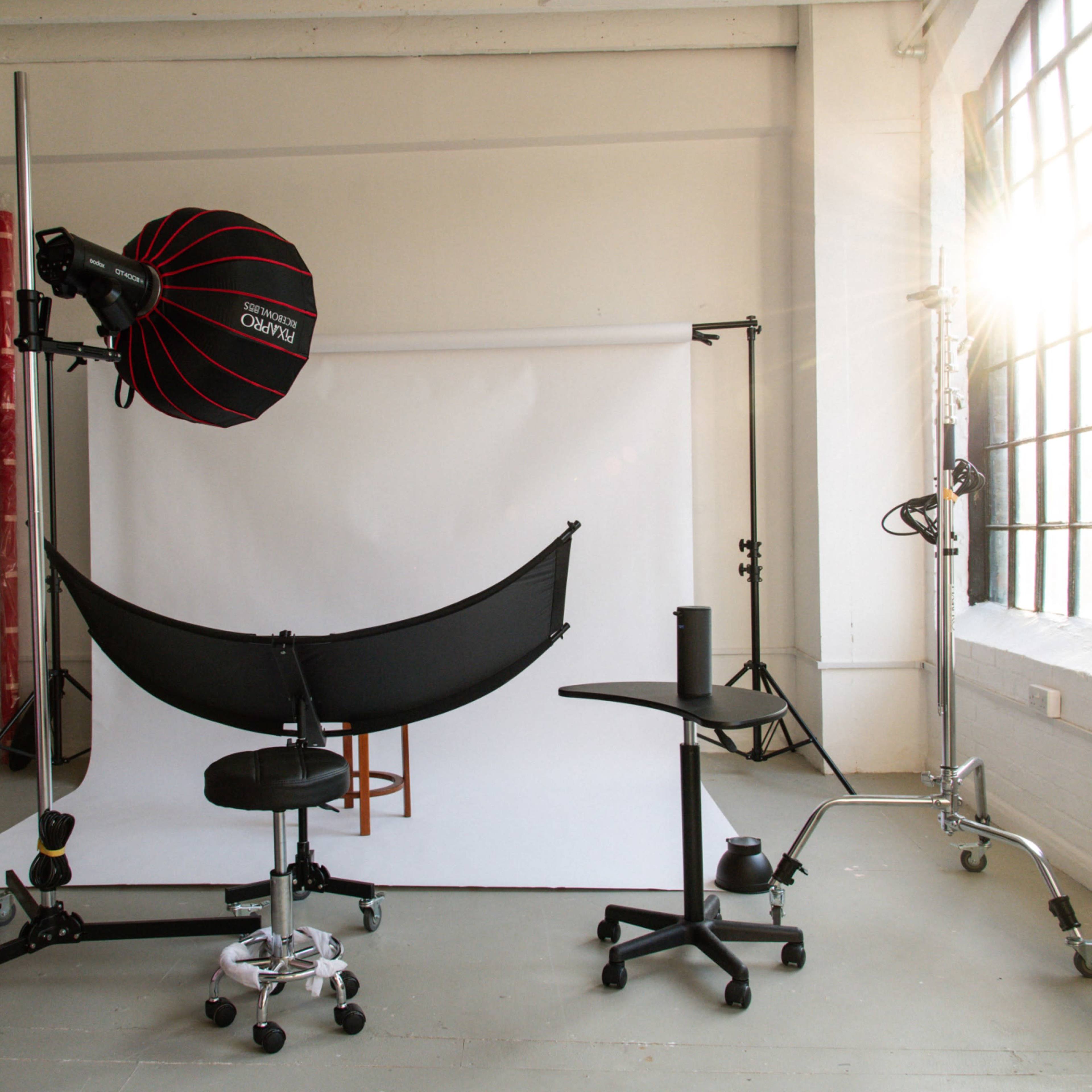 A photography studio with a backdrop, lighting equipment, and various chairs arranged in front of a large window.