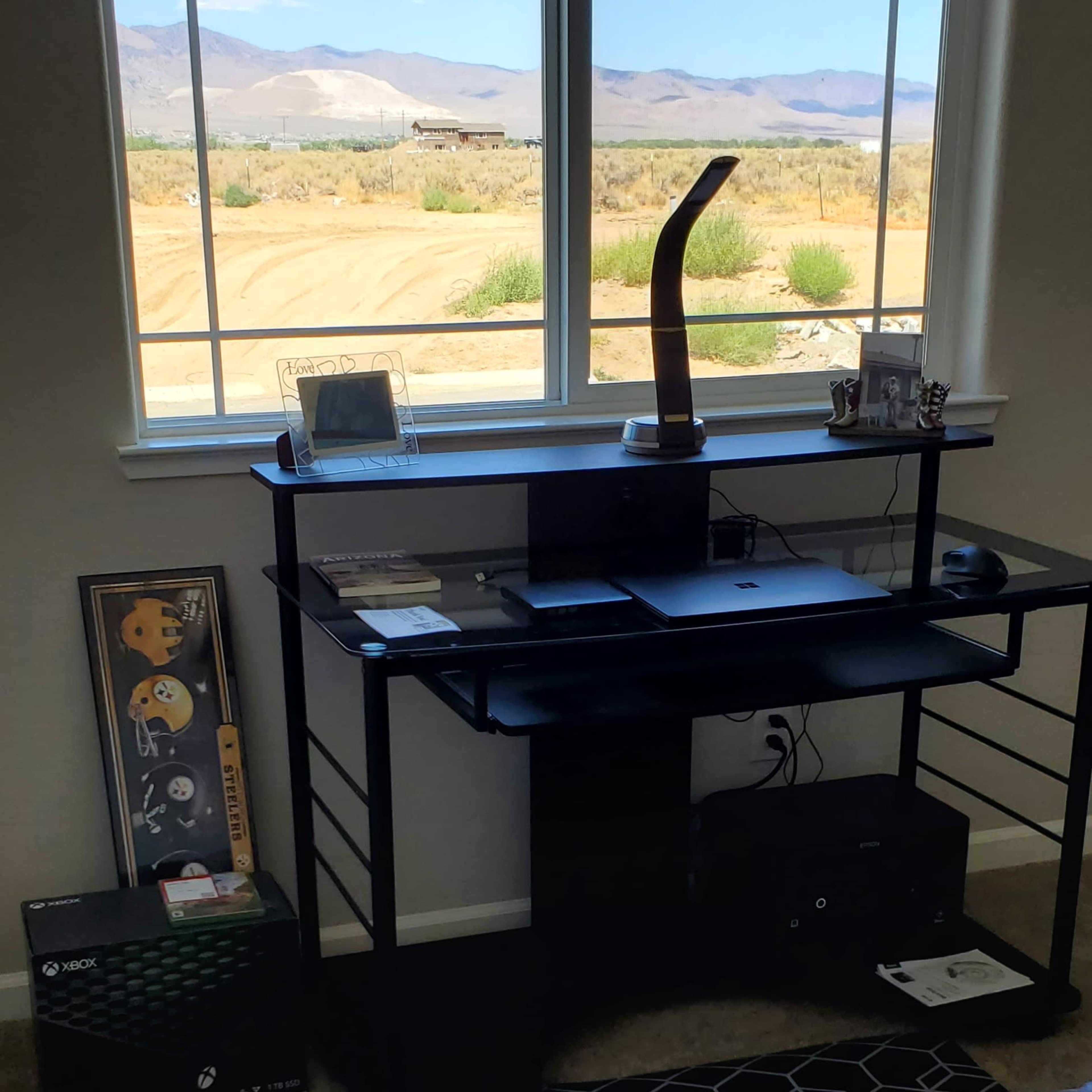 A black desk with a computer and decorative items sits near a large window overlooking a desert landscape with mountains in the distance.