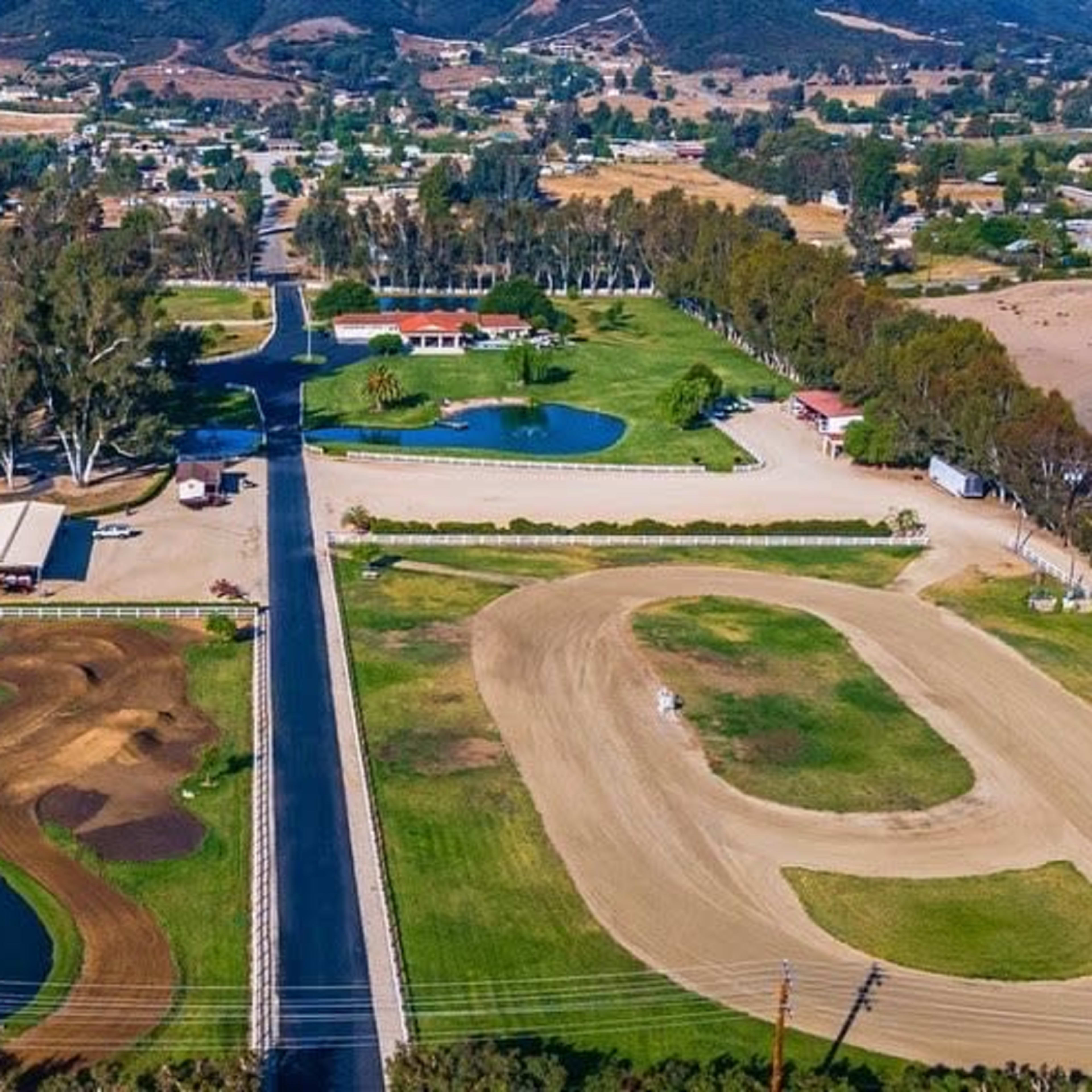 An aerial view of a rural area featuring a horse track, a motocross track, and several ponds surrounded by trees and buildings.