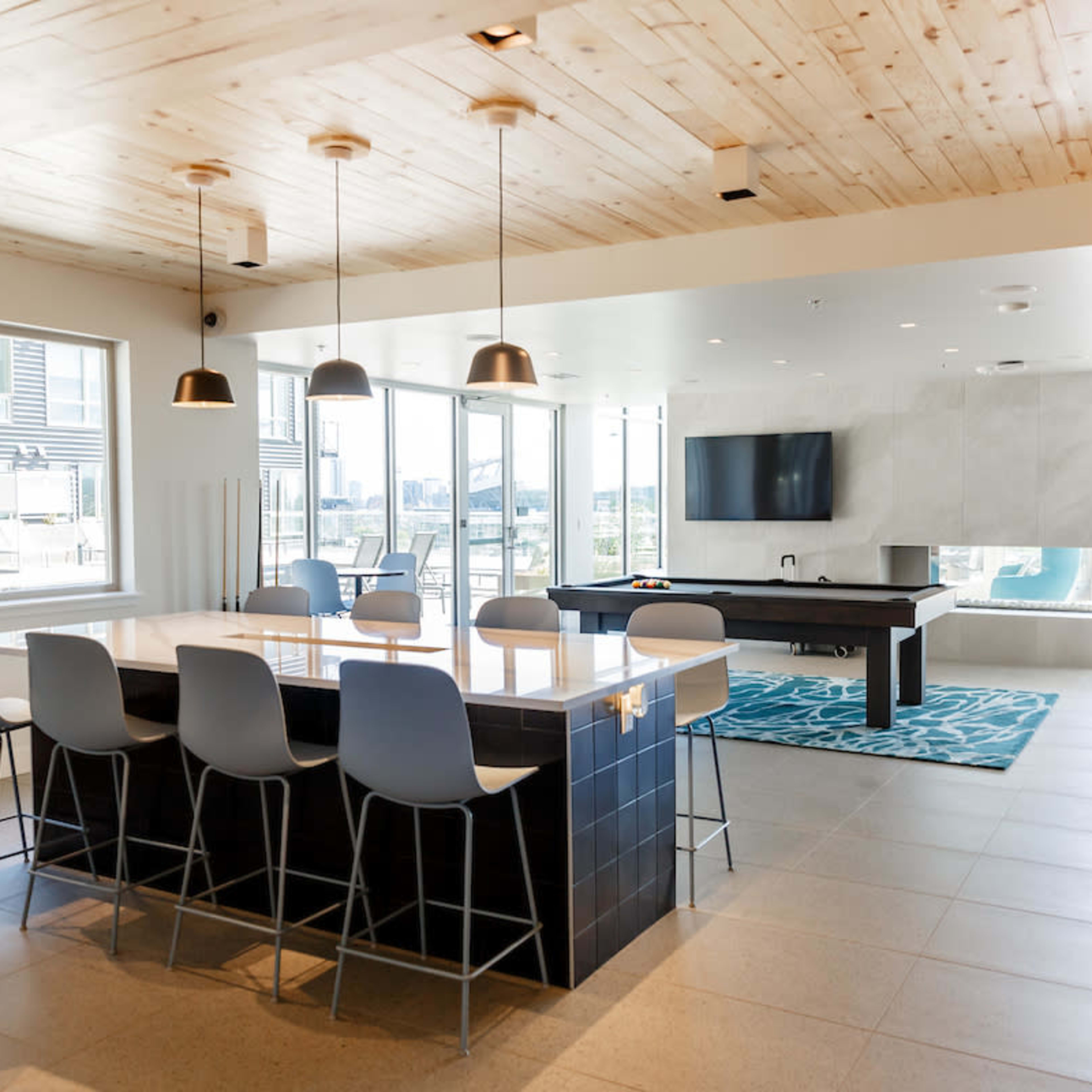 A modern interior space featuring a large island bar with seating, a living area with a television, and large windows that allow natural light.
