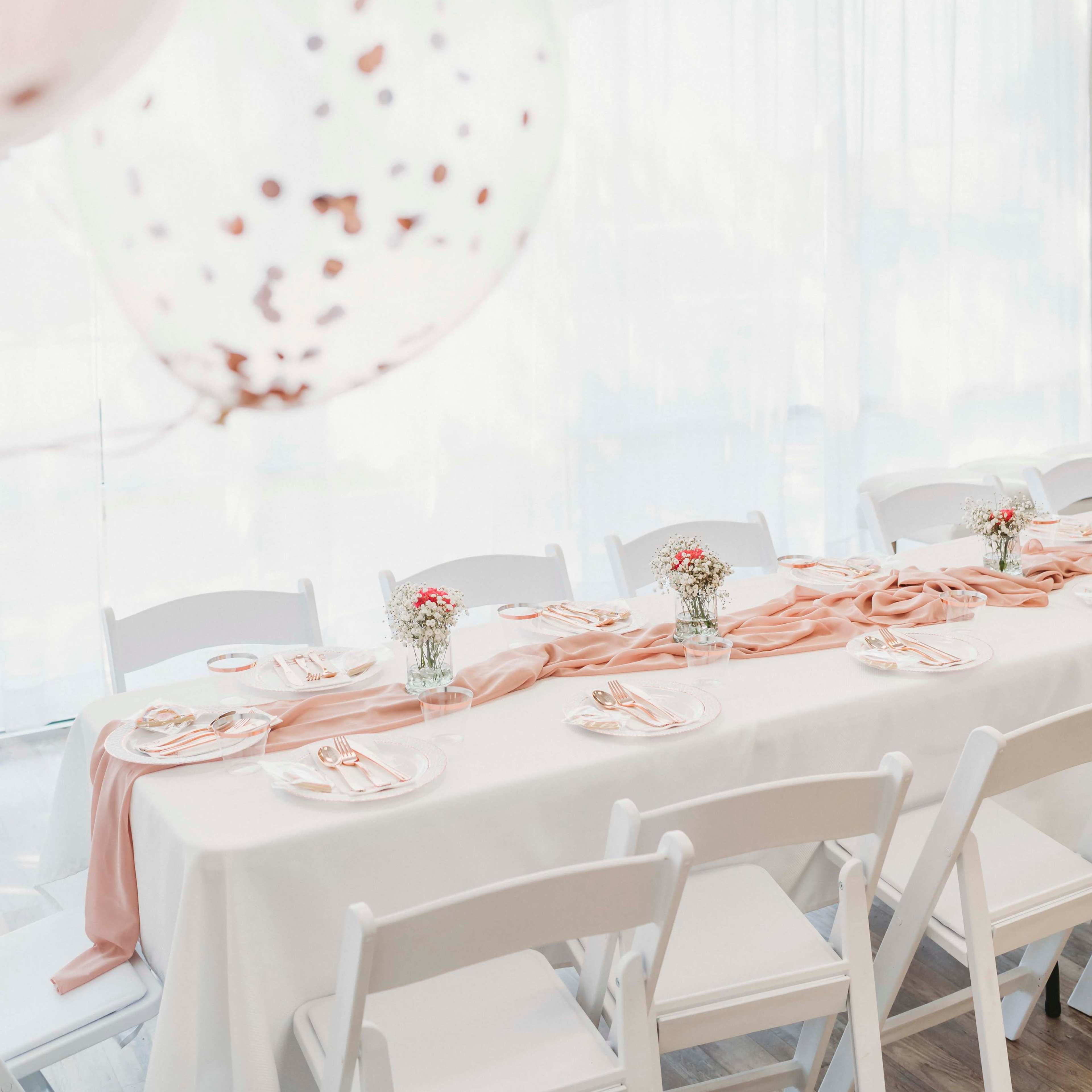 A long table is elegantly set with white tablecloths, light pink runners, and small floral centerpieces, surrounded by white chairs.