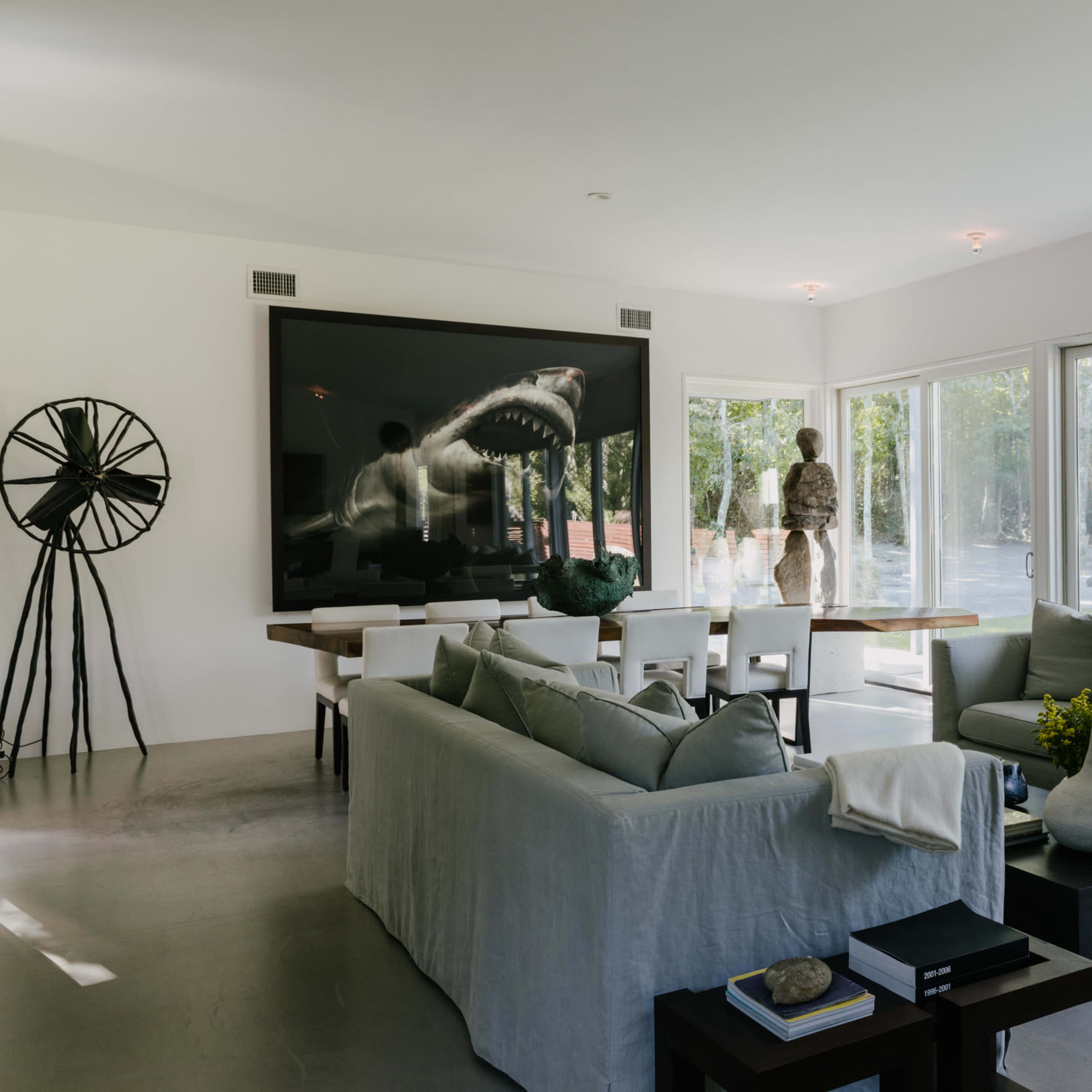 A modern living room featuring a large shark artwork, a dining table, and large glass windows overlooking a green outdoor area.