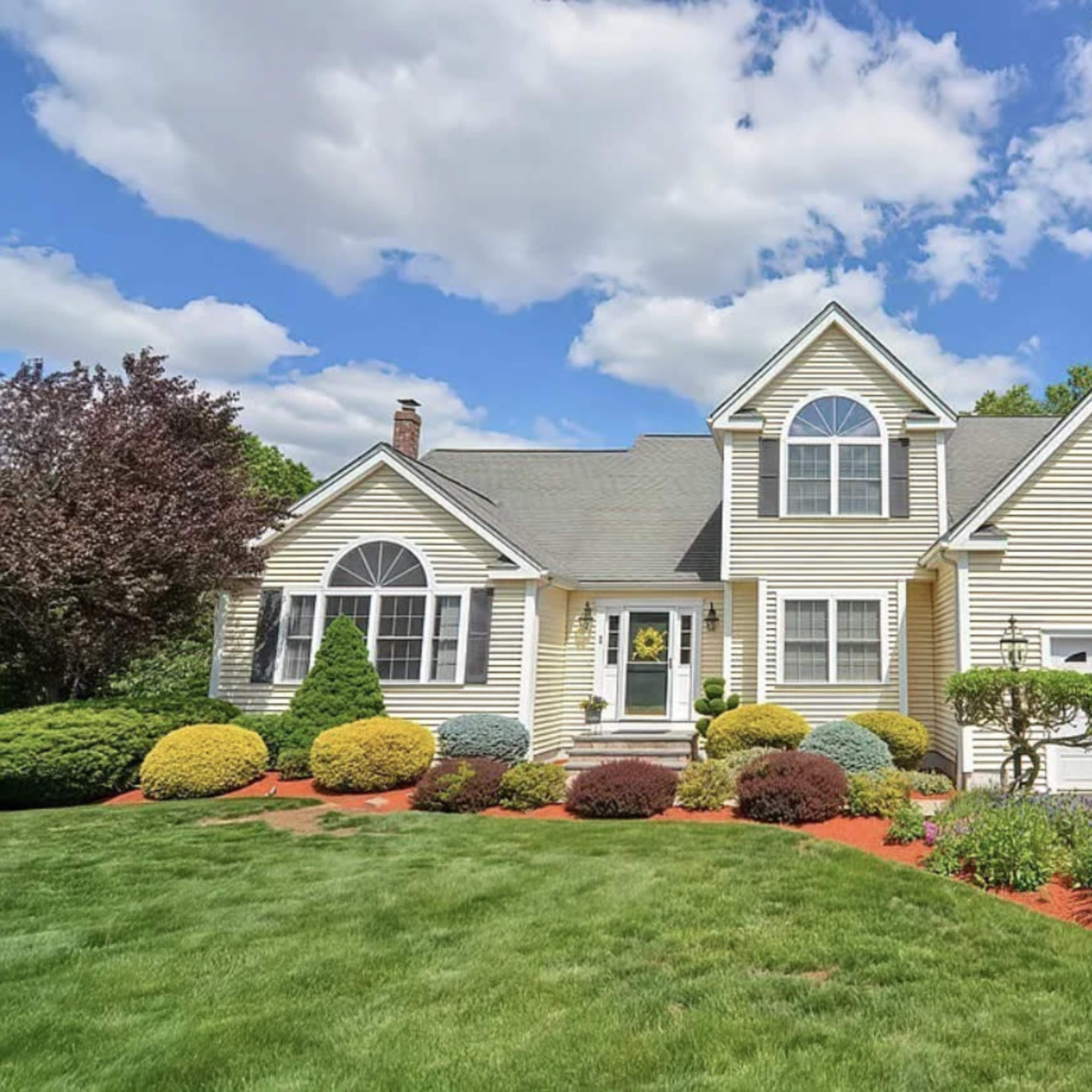 A two-story, yellow-sided house with a landscaped front yard and a driveway leading to a garage.