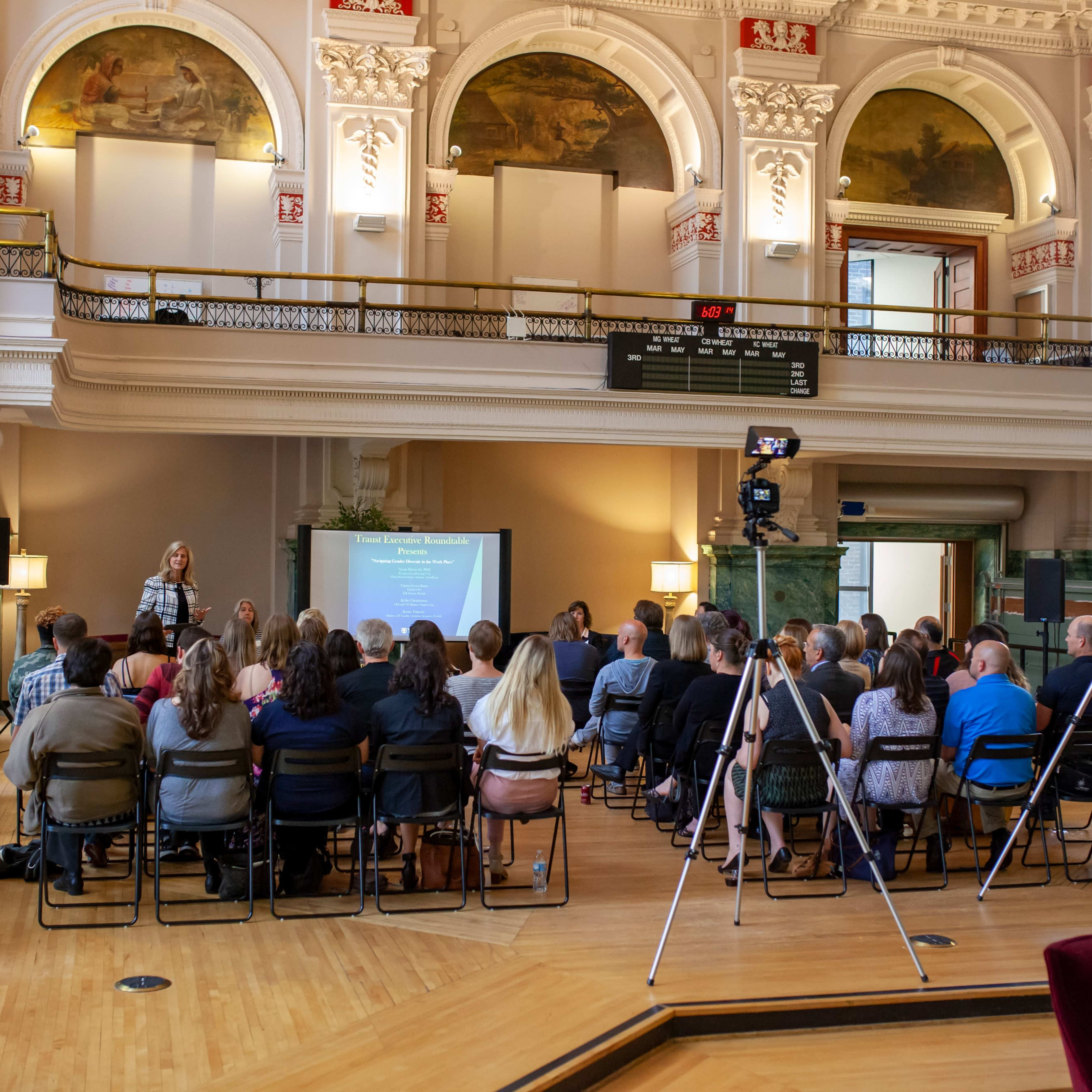 A group of individuals is seated in a hall, listening to a speaker in front of a presentation screen, with large windows and decorative architecture in the background.
