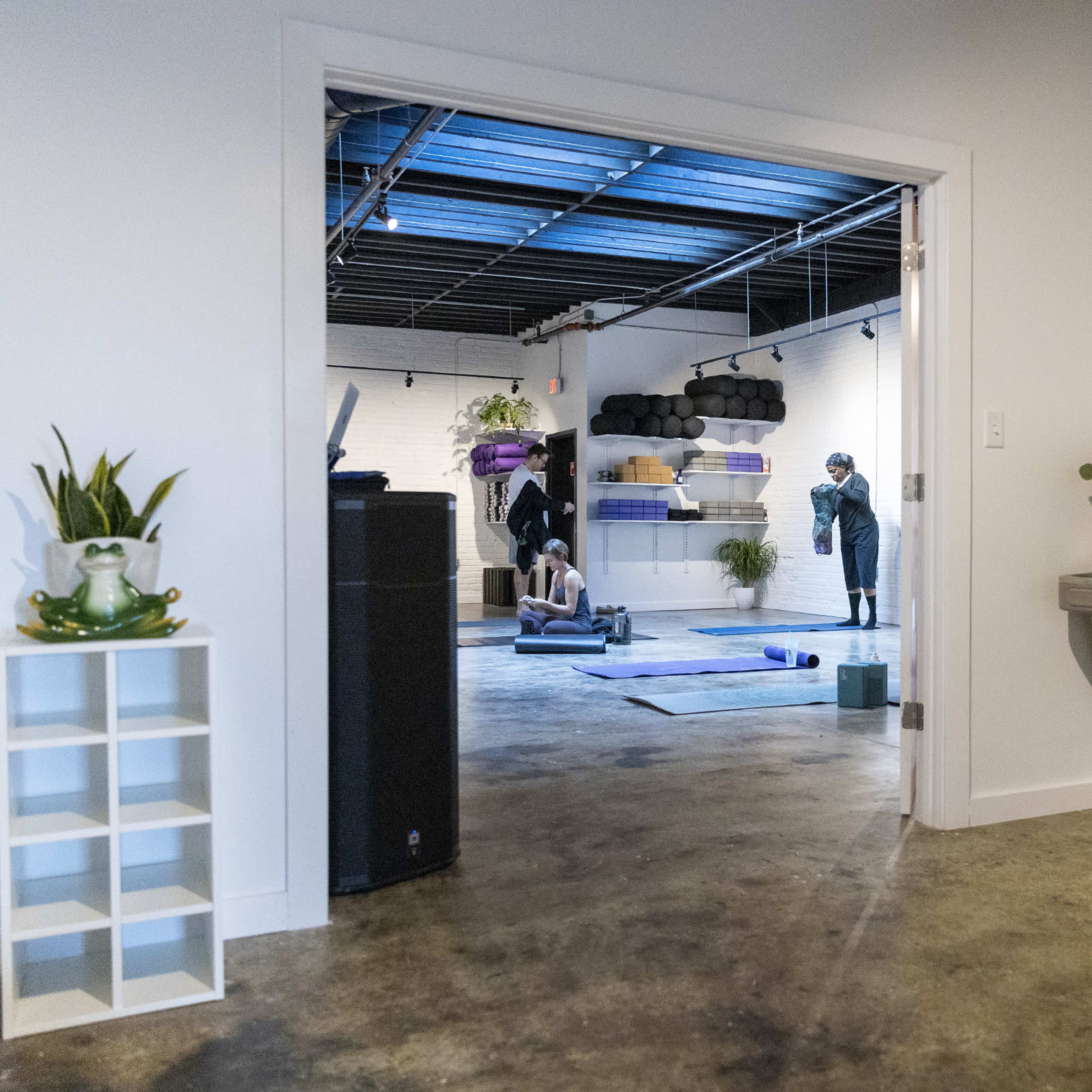 A brightly lit fitness studio features individuals practicing yoga on mats, with workout equipment and plants visible in the background.