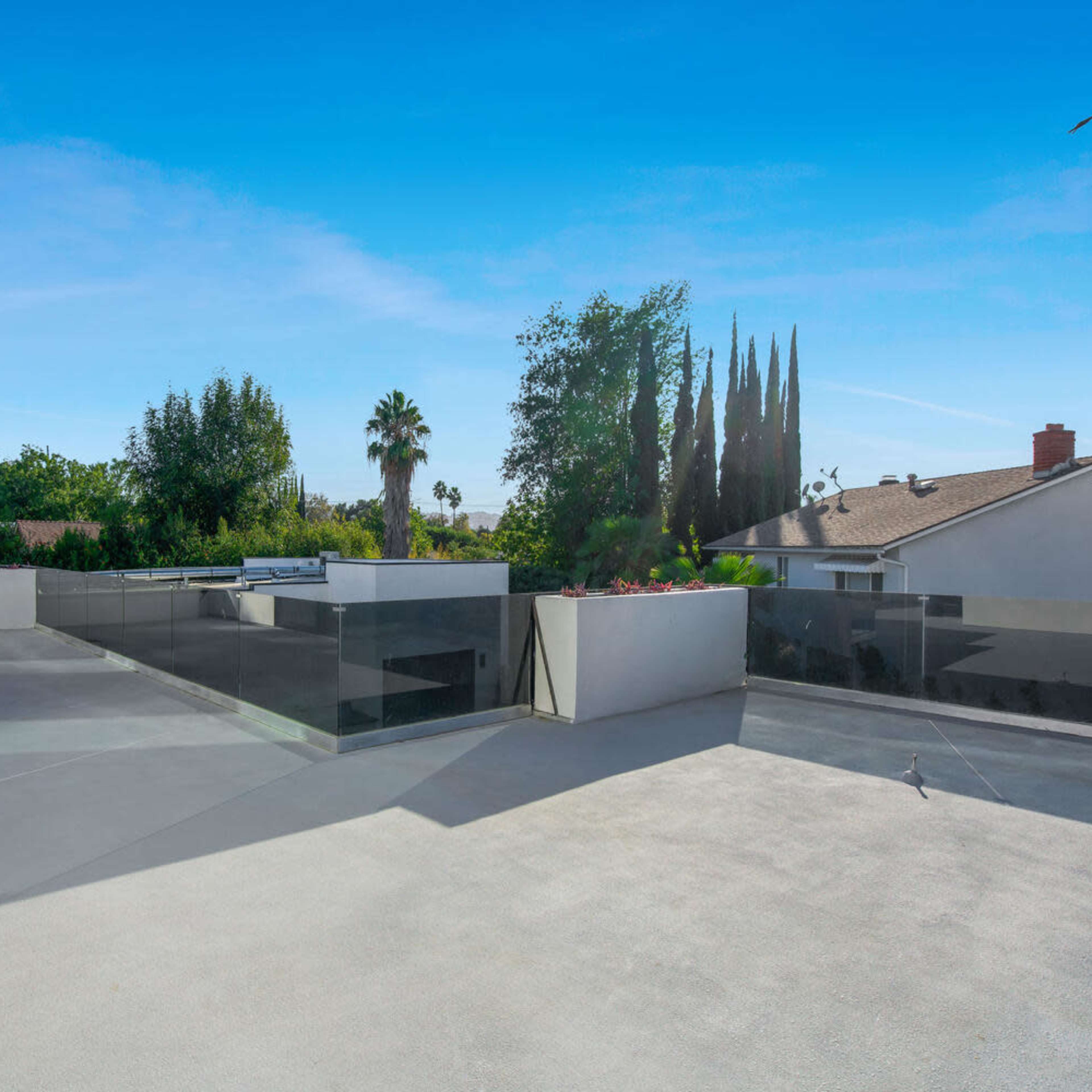 A spacious rooftop terrace features glass railings and offers a view of surrounding trees and buildings under a clear blue sky.