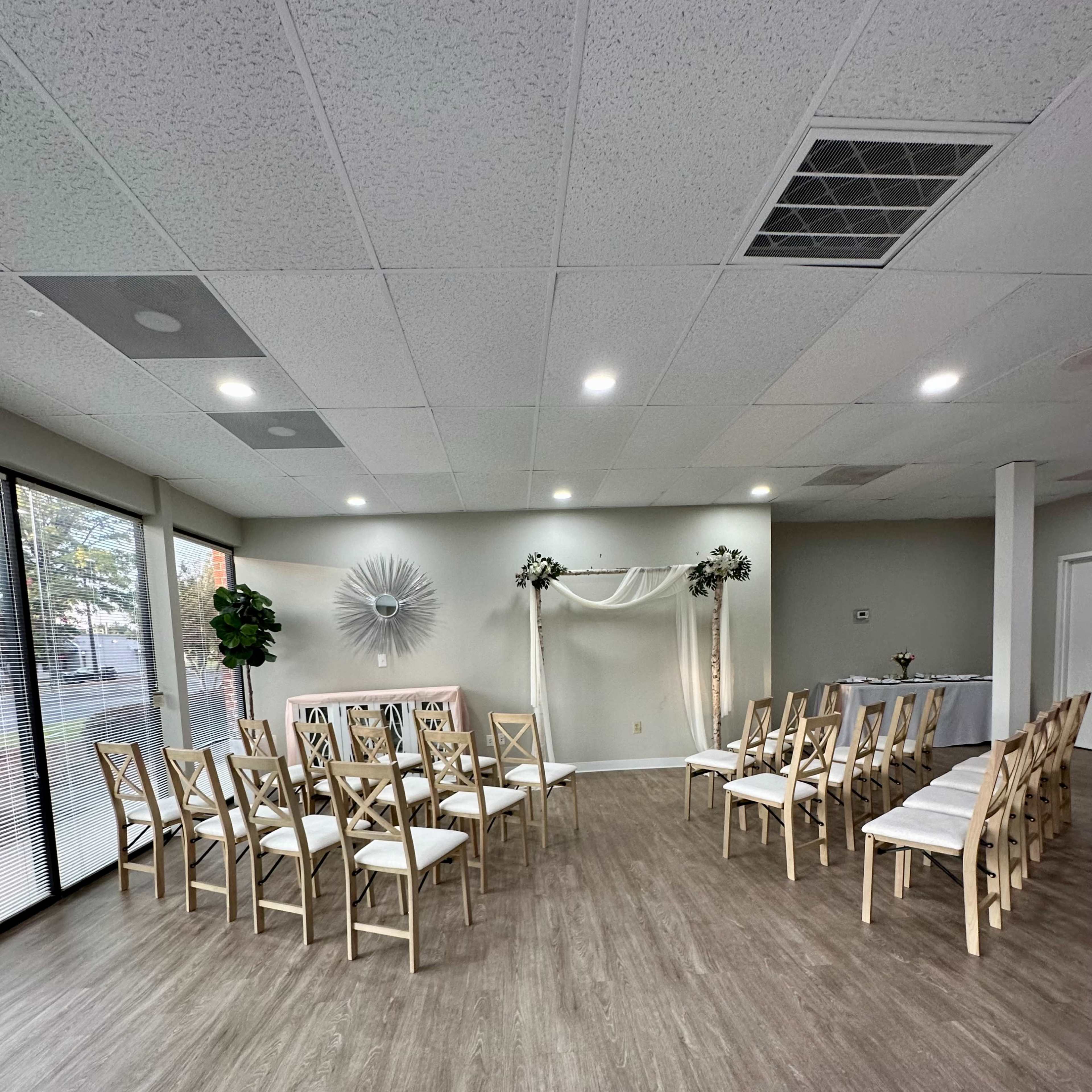 A light-filled event space set up with wooden chairs arranged in two facing rows, with a decorative arch and floral accents at the front.